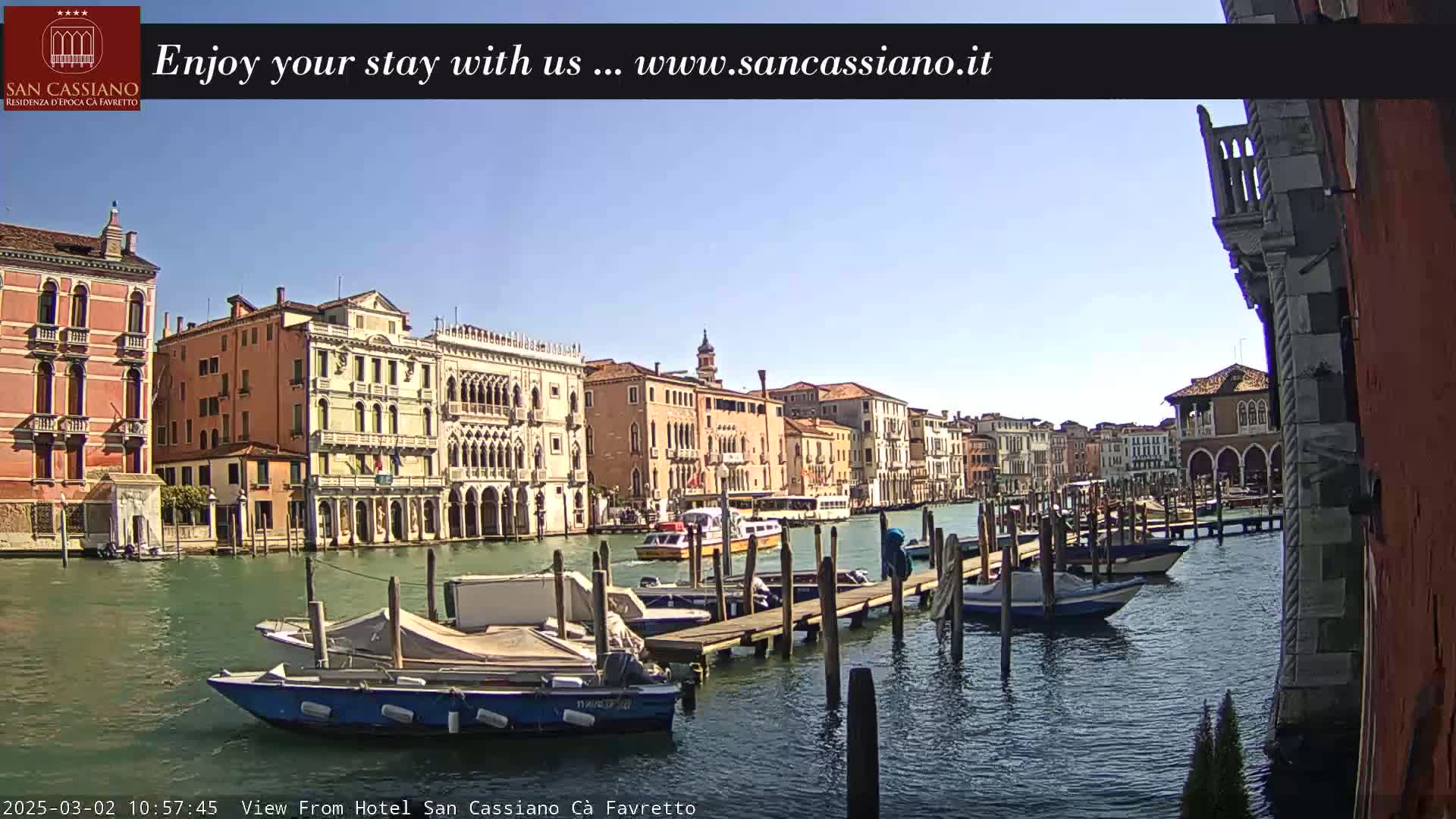 A sunny day in Venice shows several boats moored at a pier along a canal, with colorful buildings lining the waterway.