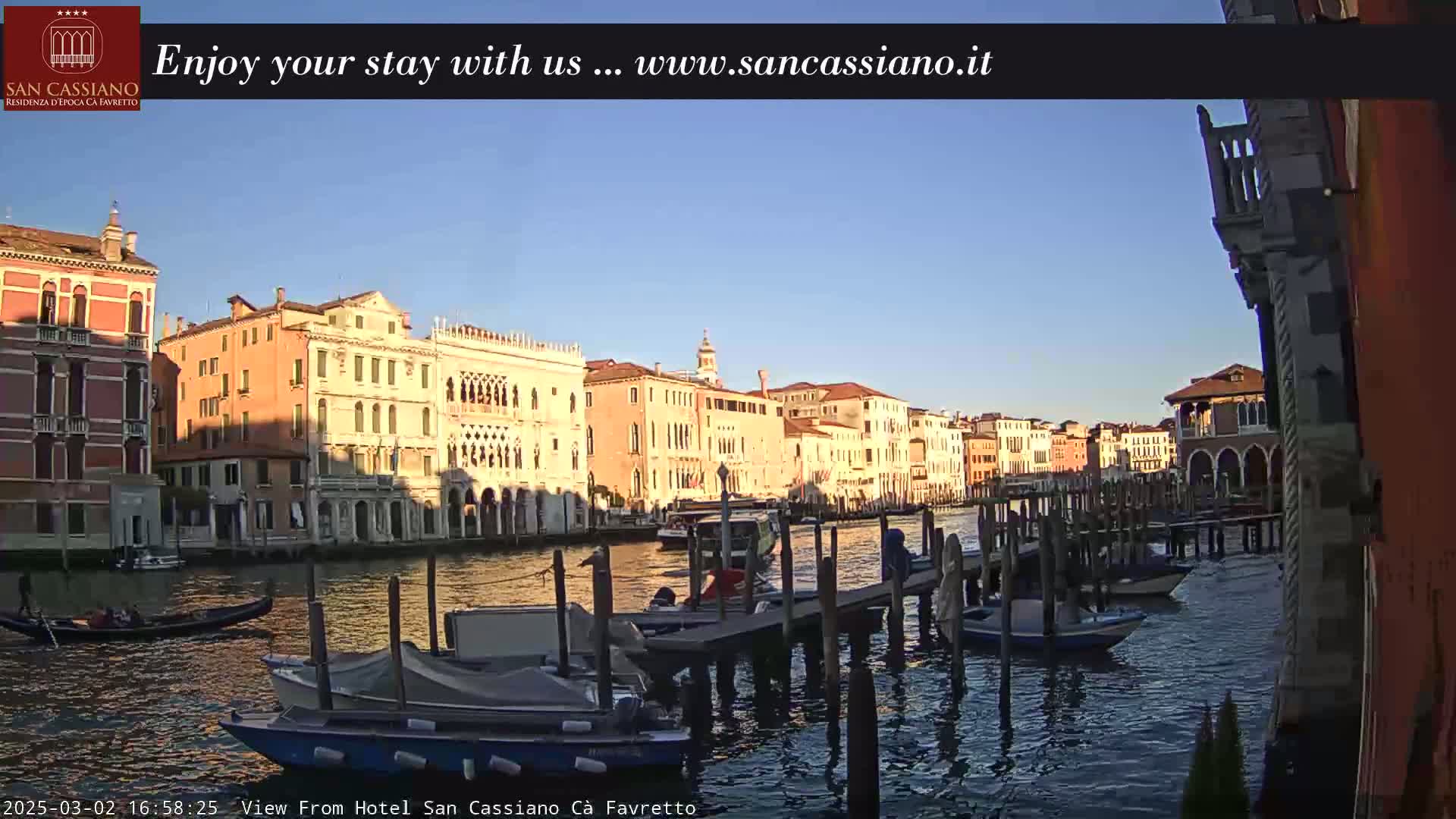 A tranquil canal scene in Venice, bathed in the warm light of a clear sunset, shows several boats moored at a dock alongside colorful buildings.