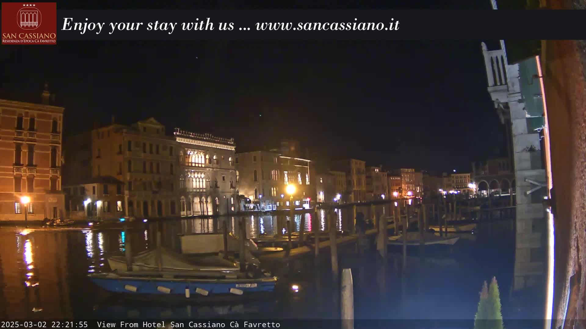 A nighttime view of a canal in Venice, Italy, shows several buildings lit up along the waterway, with boats moored along the edge.