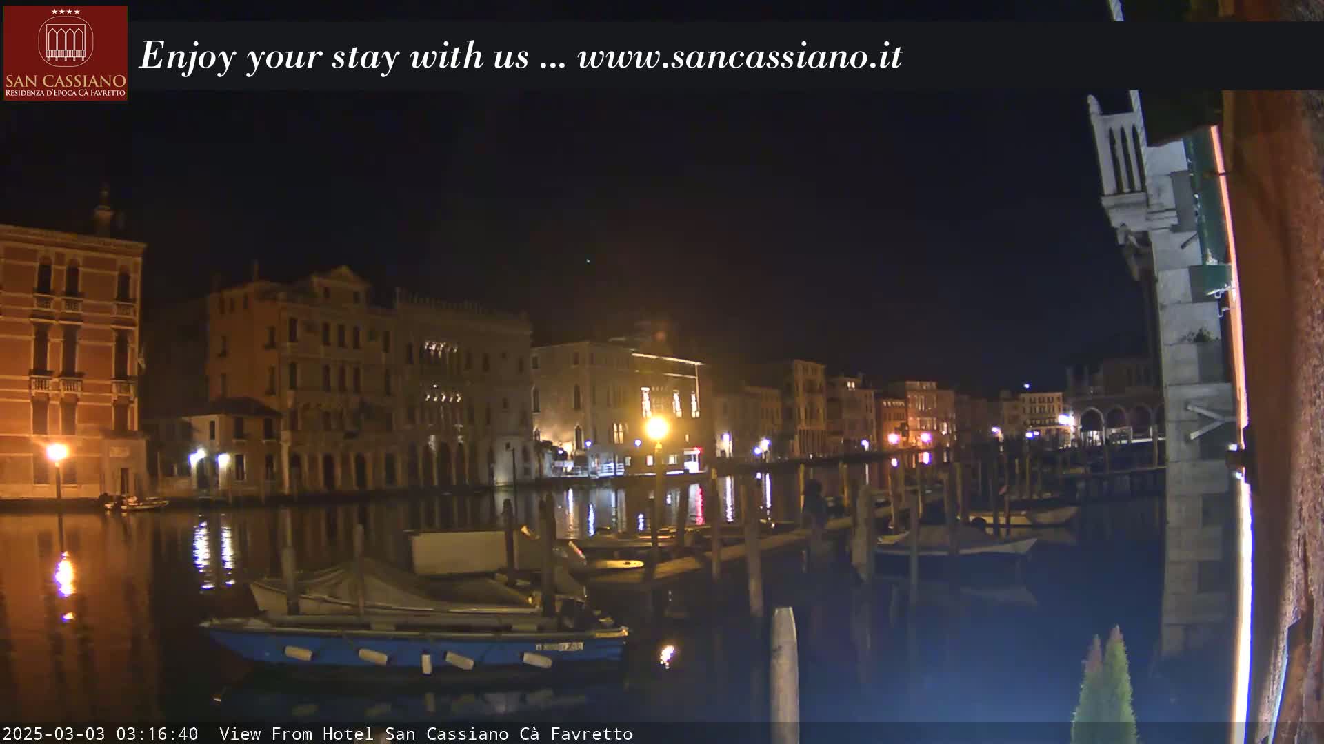 At night, illuminated buildings line a calm canal with moored boats reflecting light in the dark water.