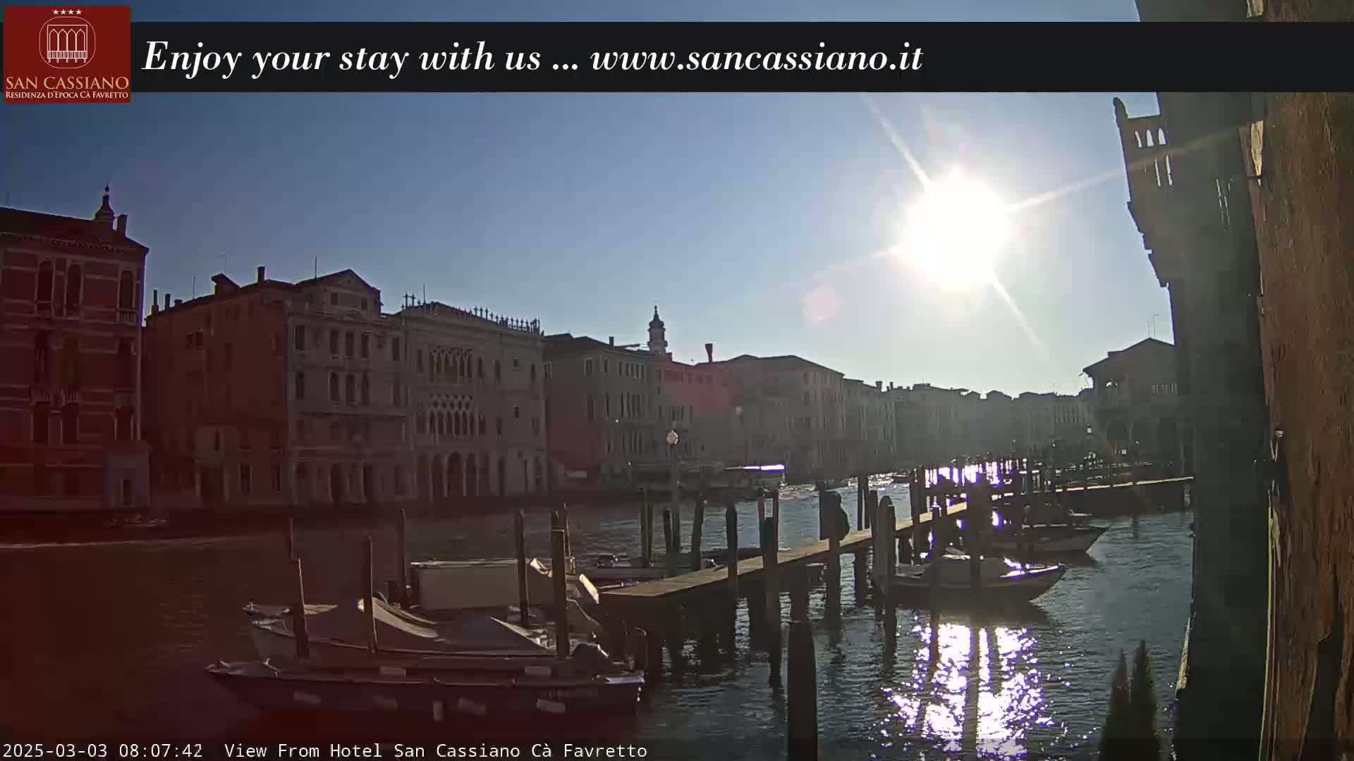 A sunny day in Venice shows a canal with boats moored at wooden docks, lined by historic buildings.