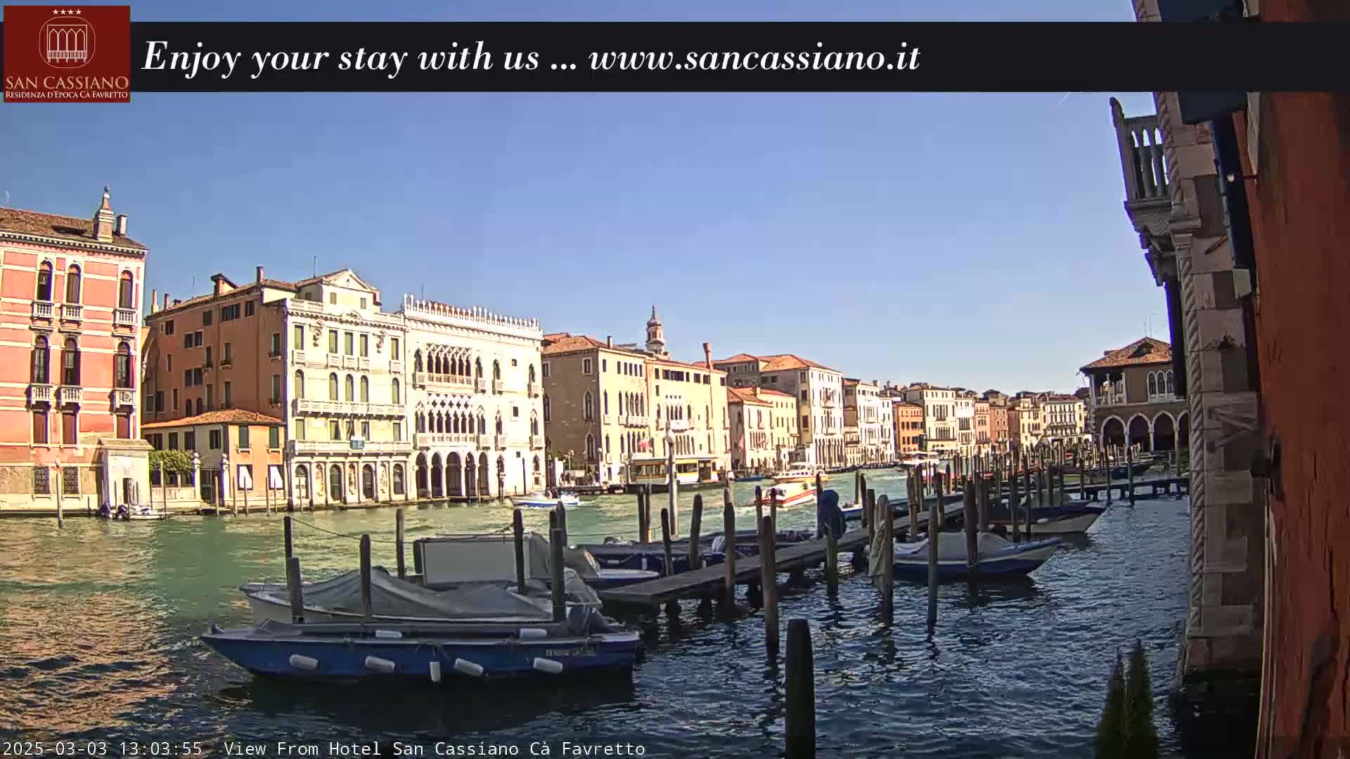 A sunny day in Venice shows several canal-side buildings and moored boats along a waterway.
