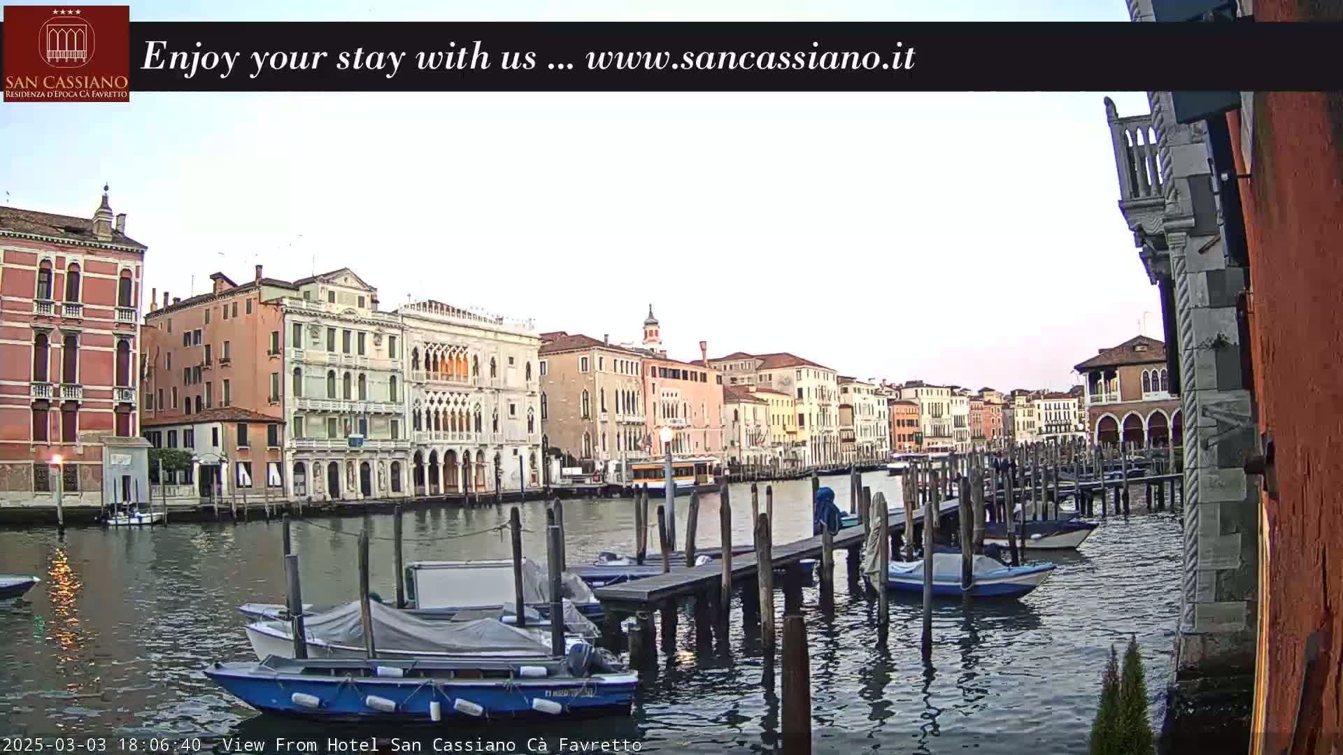 A calm canal in Venice, Italy, is lined with colorful buildings at dusk, and several boats are moored at a dock.