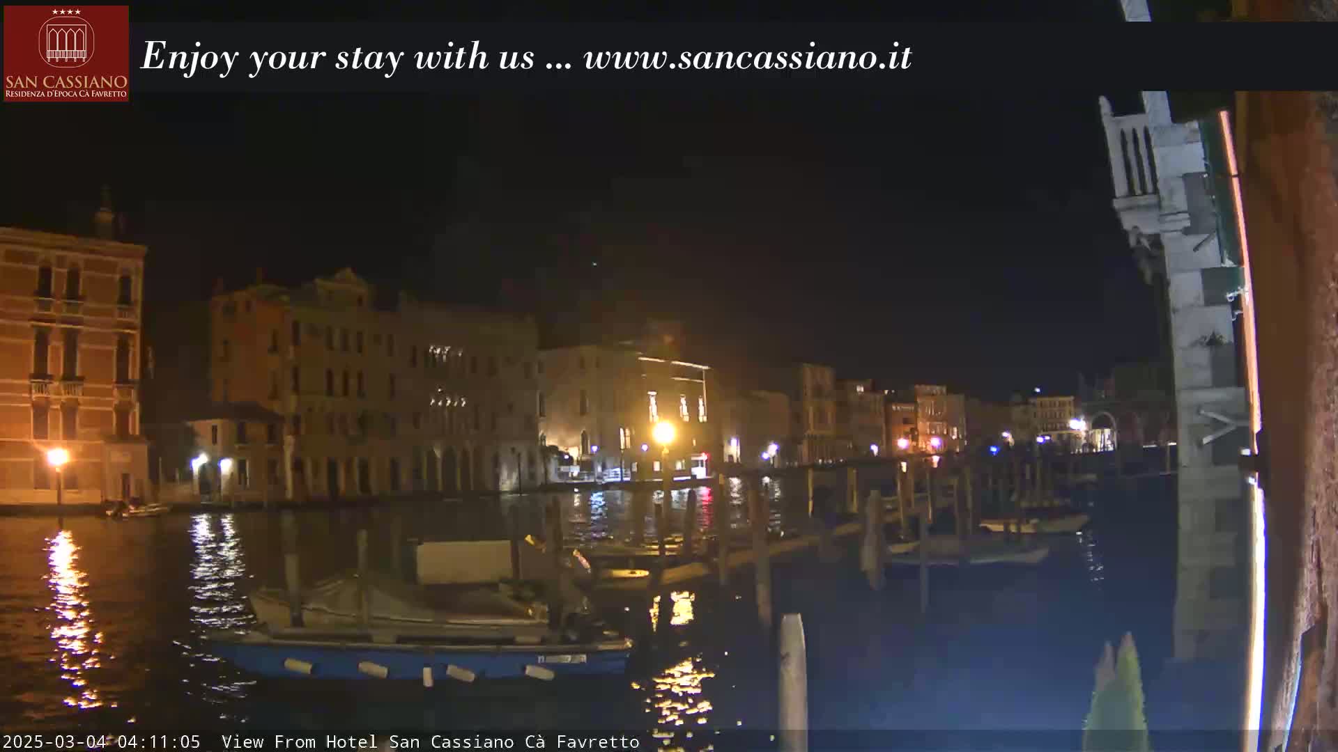 At night, a canal in Venice is shown with several buildings lit up along the waterway, and boats moored at a dock.