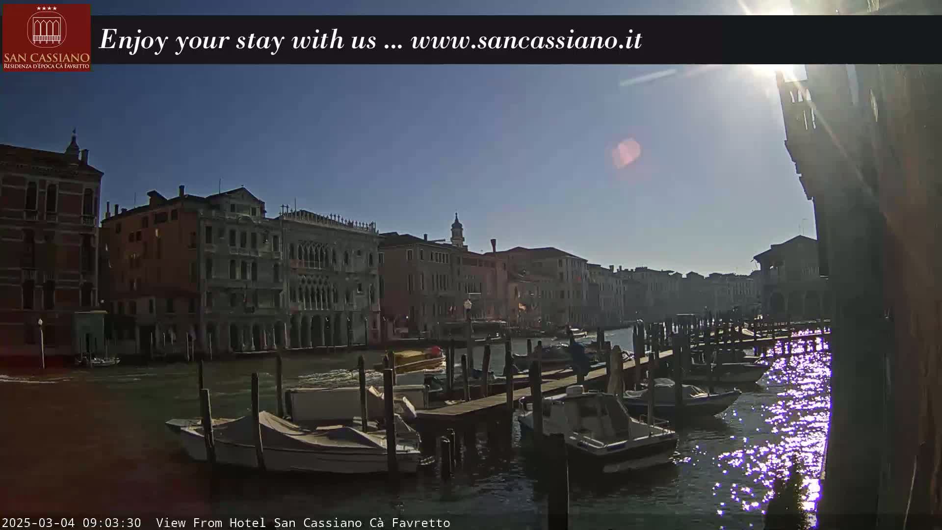 A sunny day in Venice shows a canal lined with buildings and several boats moored at a dock.
