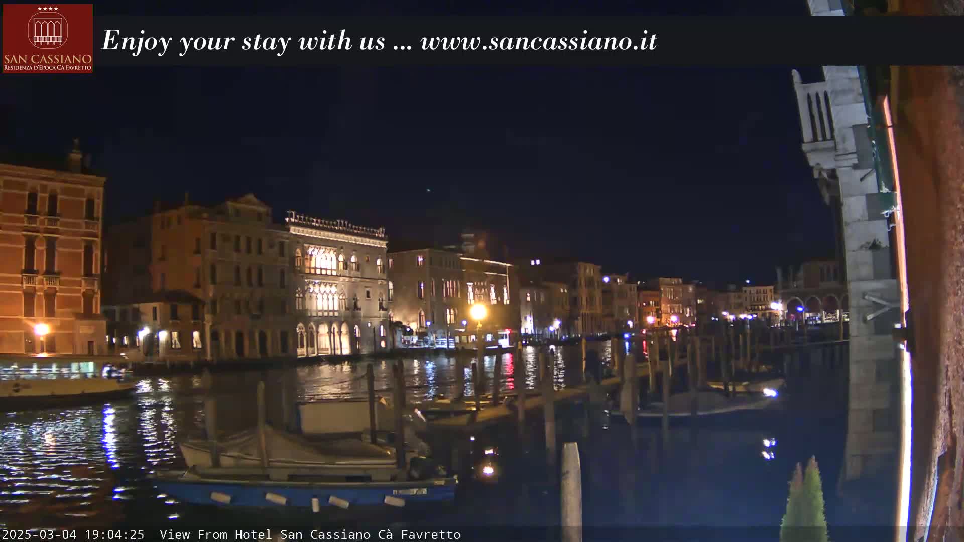 A nighttime view of a canal in Venice, Italy, shows illuminated buildings lining the waterway, with boats moored along the edges.