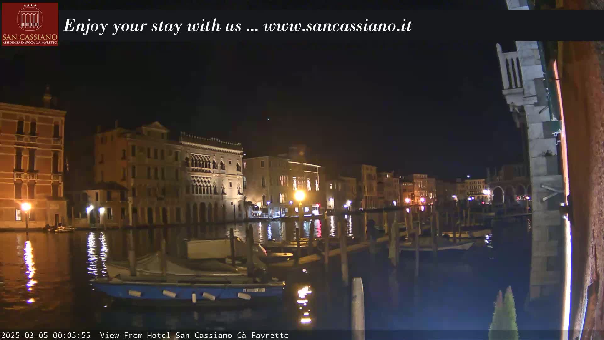 A nighttime view of a canal in Venice, Italy, showing illuminated buildings along the waterway and several boats moored at the edge.