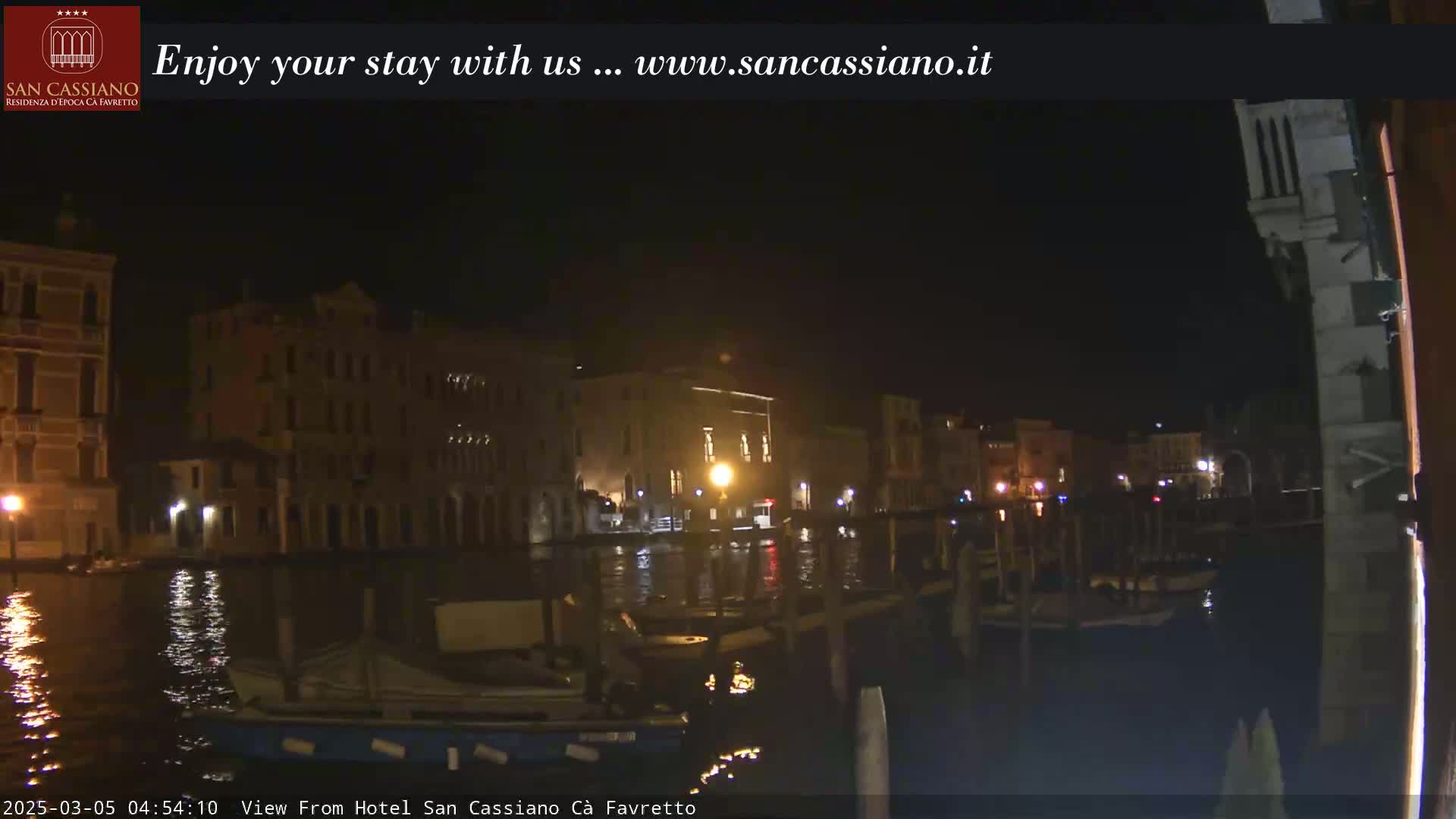 A nighttime view of a canal in Venice, Italy, shows several buildings lit up along the waterway, with boats moored along the canal edge.