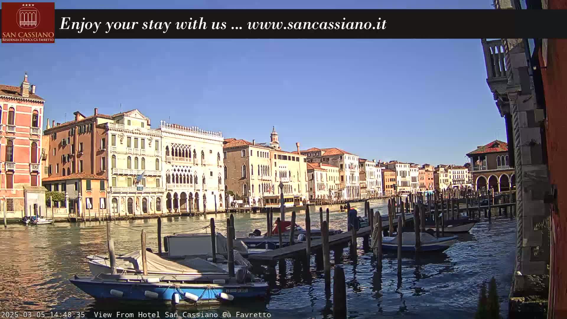 A sunny day in Venice shows a canal lined with colorful buildings and several boats moored at wooden docks.