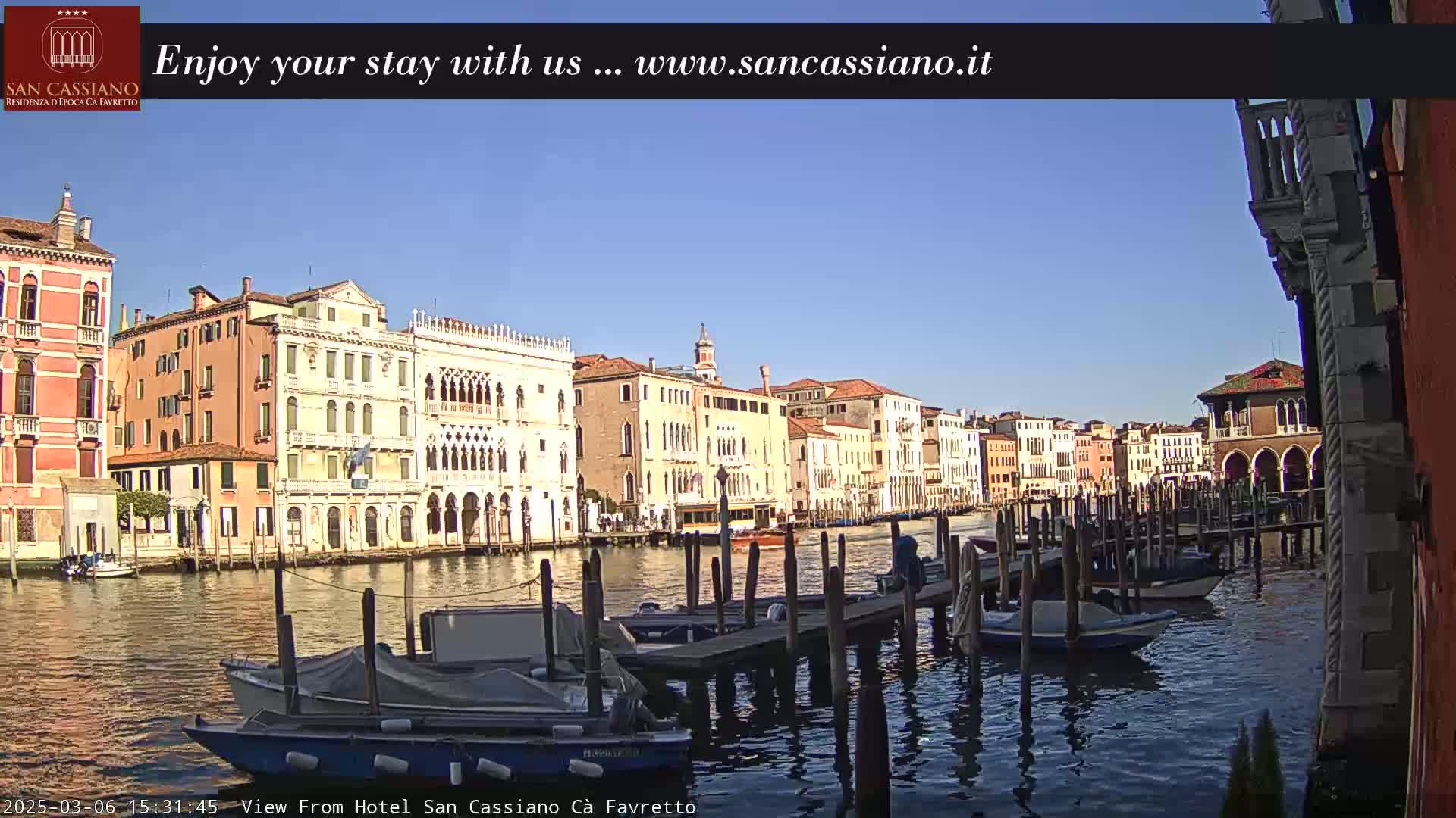 A sunny day in Venice shows a canal lined with colorful buildings and several boats moored at a dock.