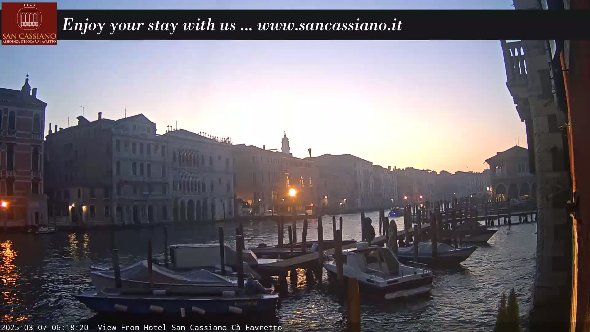 A tranquil sunrise illuminates a Venetian canal, where several motorboats are moored along a wooden dock in front of historic buildings.