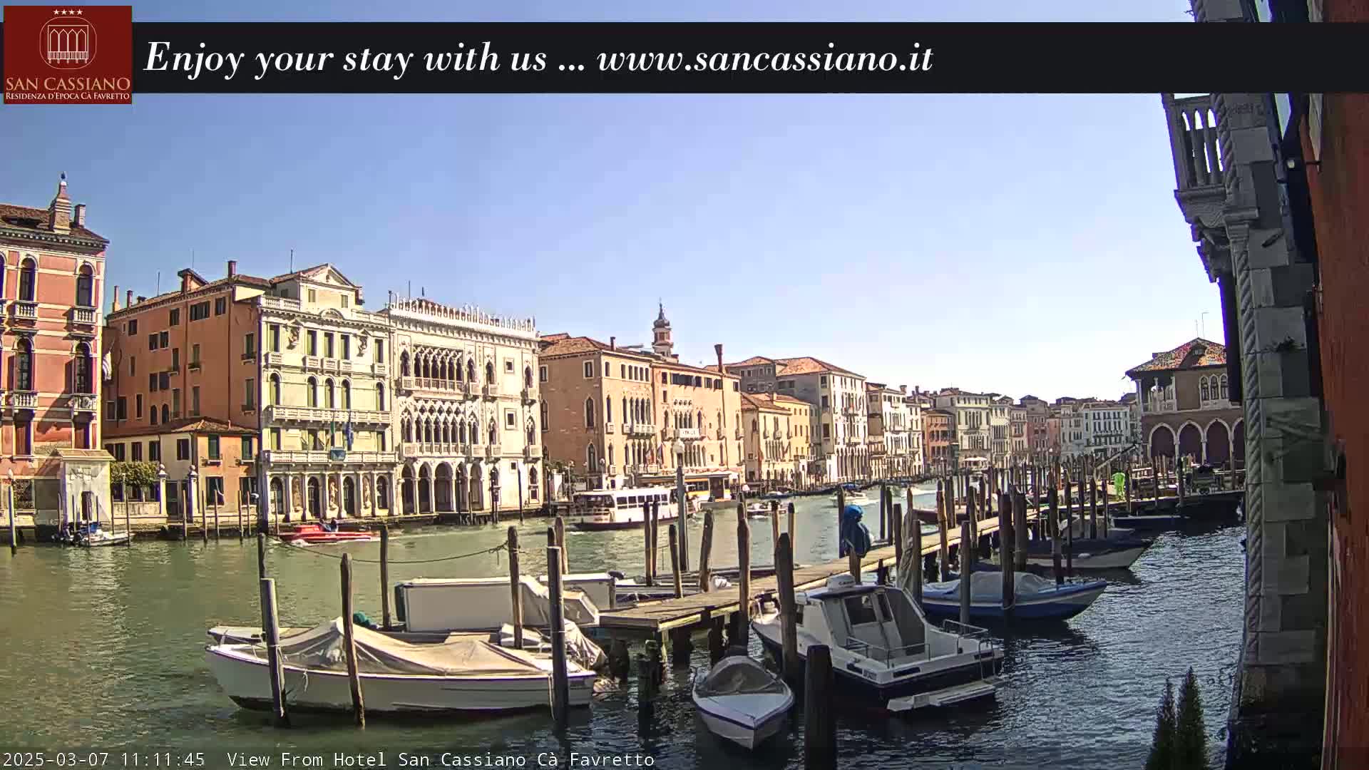 A sunny day in Venice shows a canal lined with colorful buildings and numerous boats docked at wooden piers.