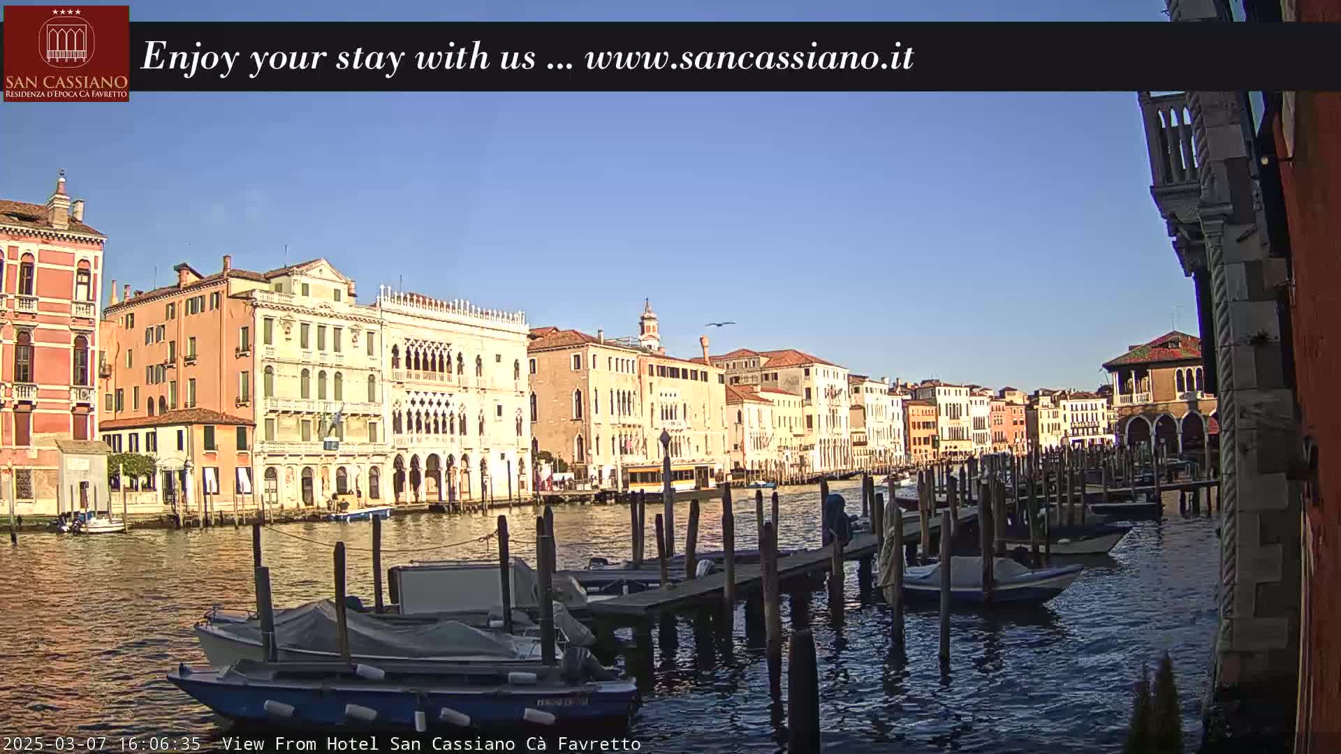 A sunny day in Venice shows several boats moored at a dock along a canal with colorful buildings lining the waterway.