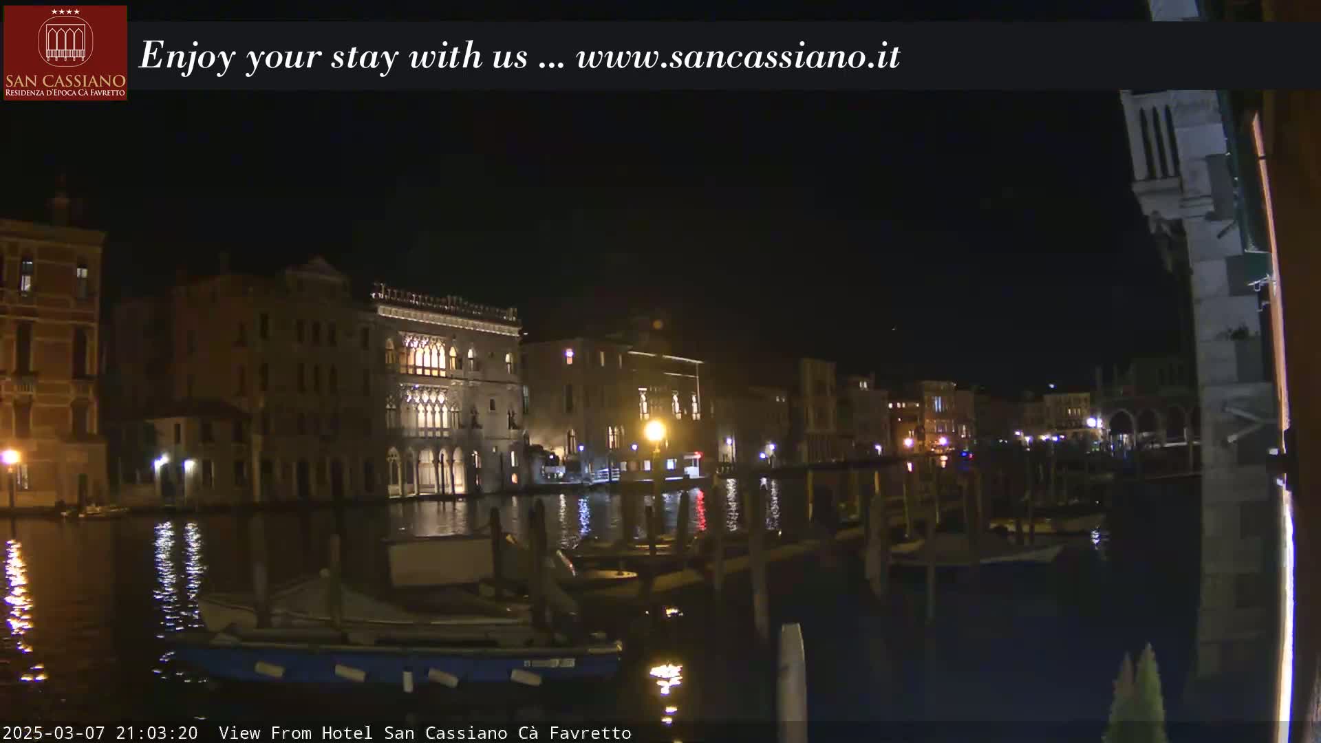 A nighttime view across a calm canal shows illuminated buildings lining the waterfront, with several boats moored along the edge.