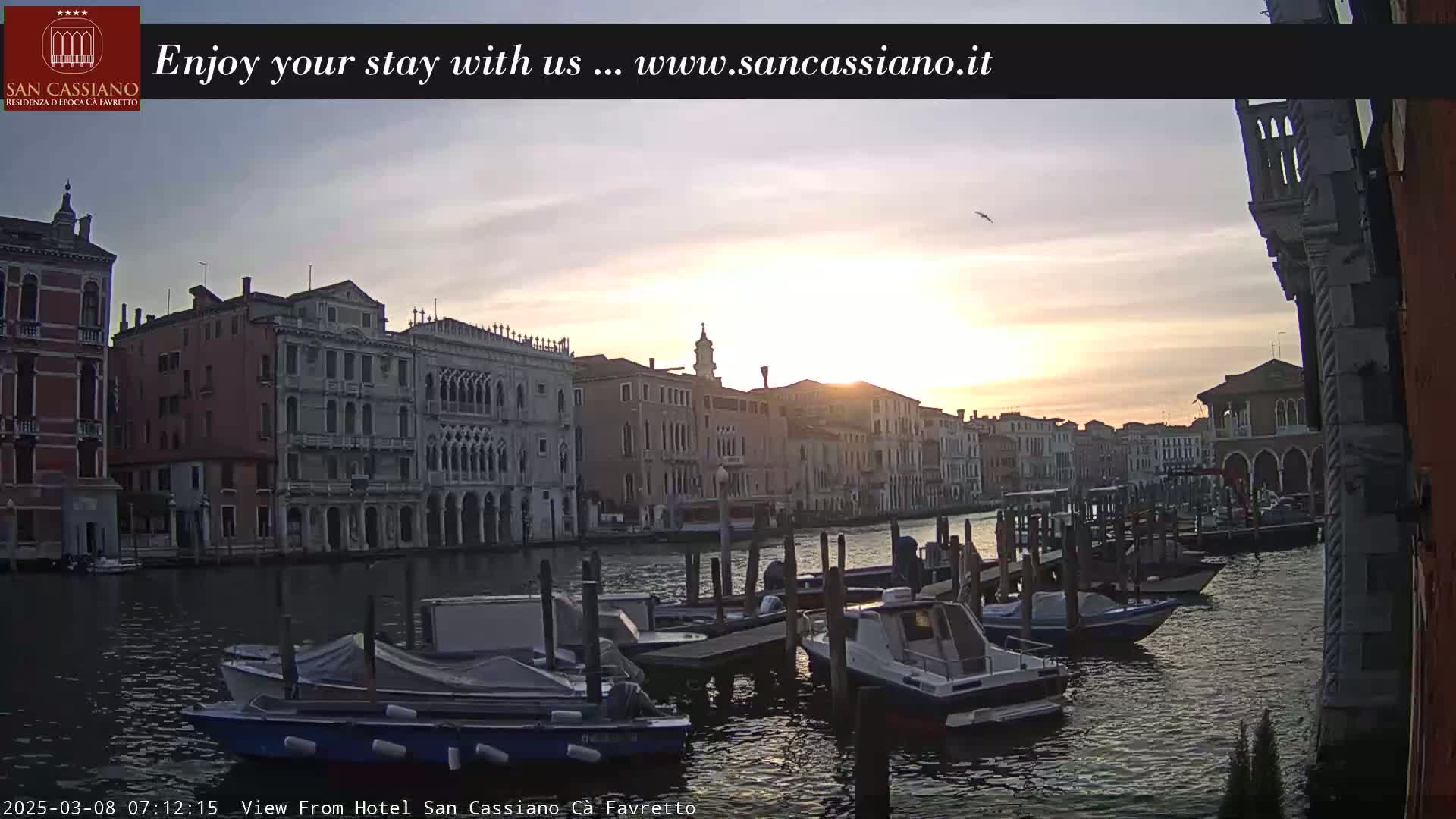 A sunrise casts a golden glow on a Venetian canal lined with historic buildings and numerous boats moored at the water's edge.