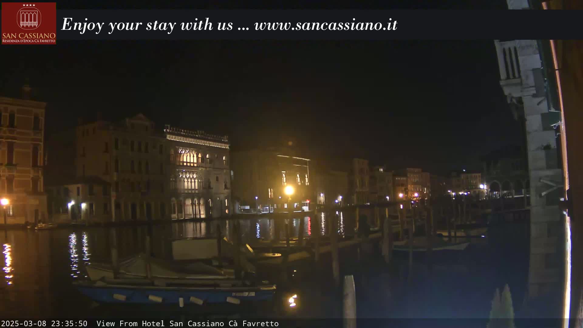 A nighttime view across a calm canal shows several illuminated buildings along the waterfront.