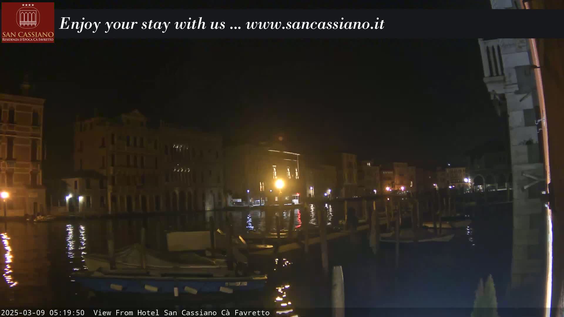 A nighttime view of a canal in Venice, with illuminated buildings lining the waterway and several boats moored along the banks.