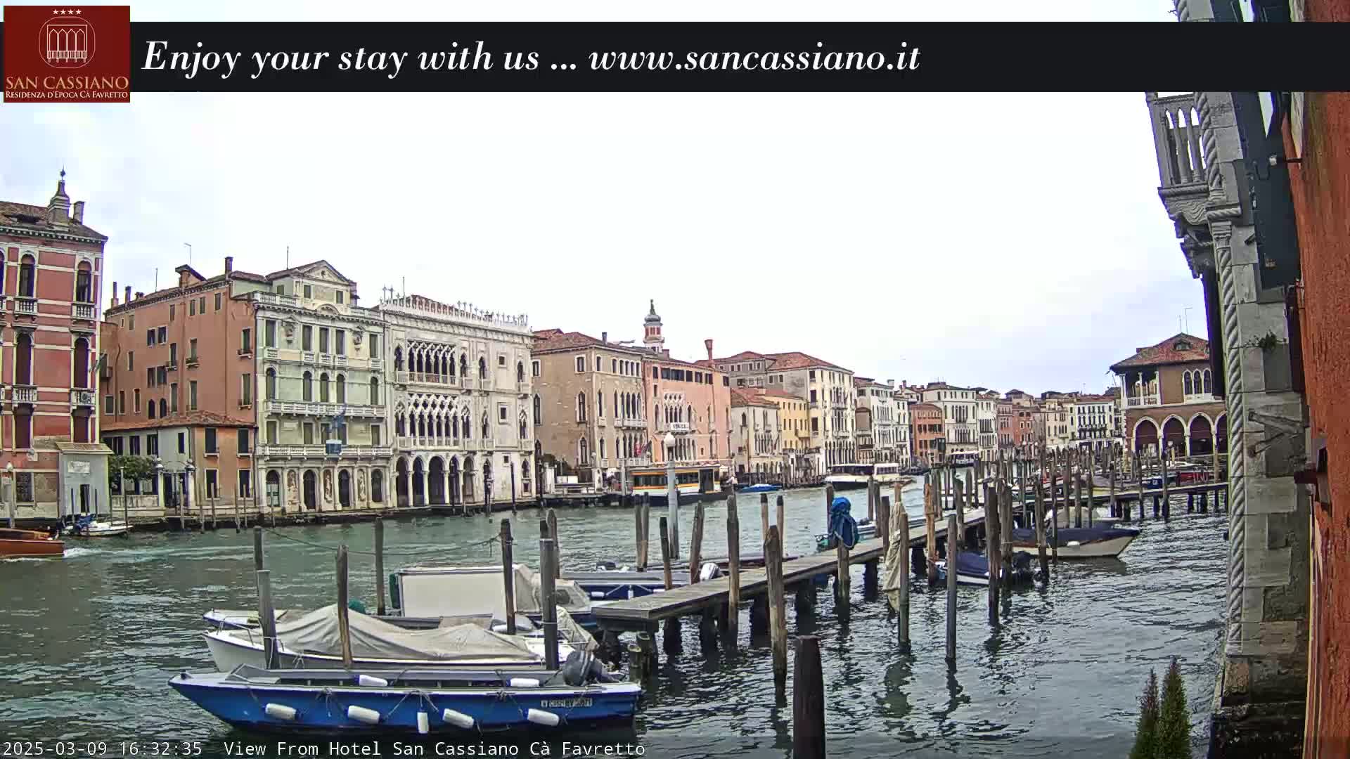 A canal in Venice, Italy, with several boats moored at wooden docks, is viewed on an overcast day, showing a line of buildings along the waterway.