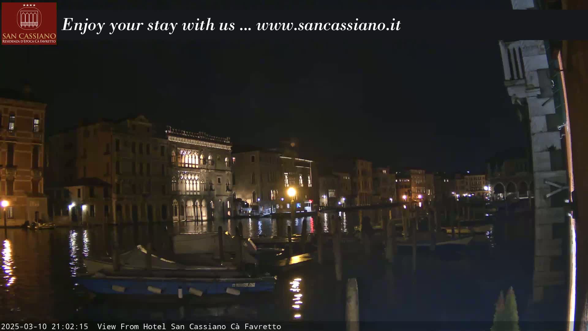 Nighttime view of illuminated buildings along a canal, with several boats moored along the edge.