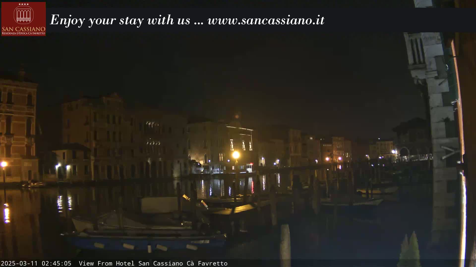 A nighttime view of a canal in Venice, Italy, shows buildings lining the waterway with boats moored along the banks under a dark sky.