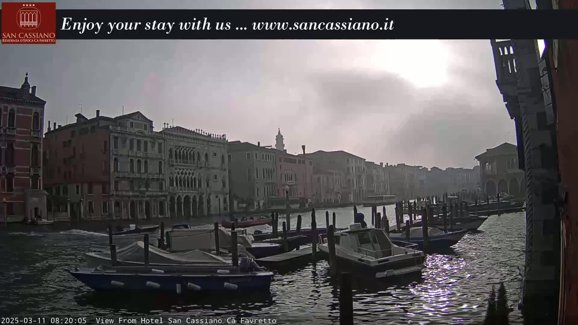 A hazy sunrise illuminates a Venetian canal lined with historic buildings and moored boats.