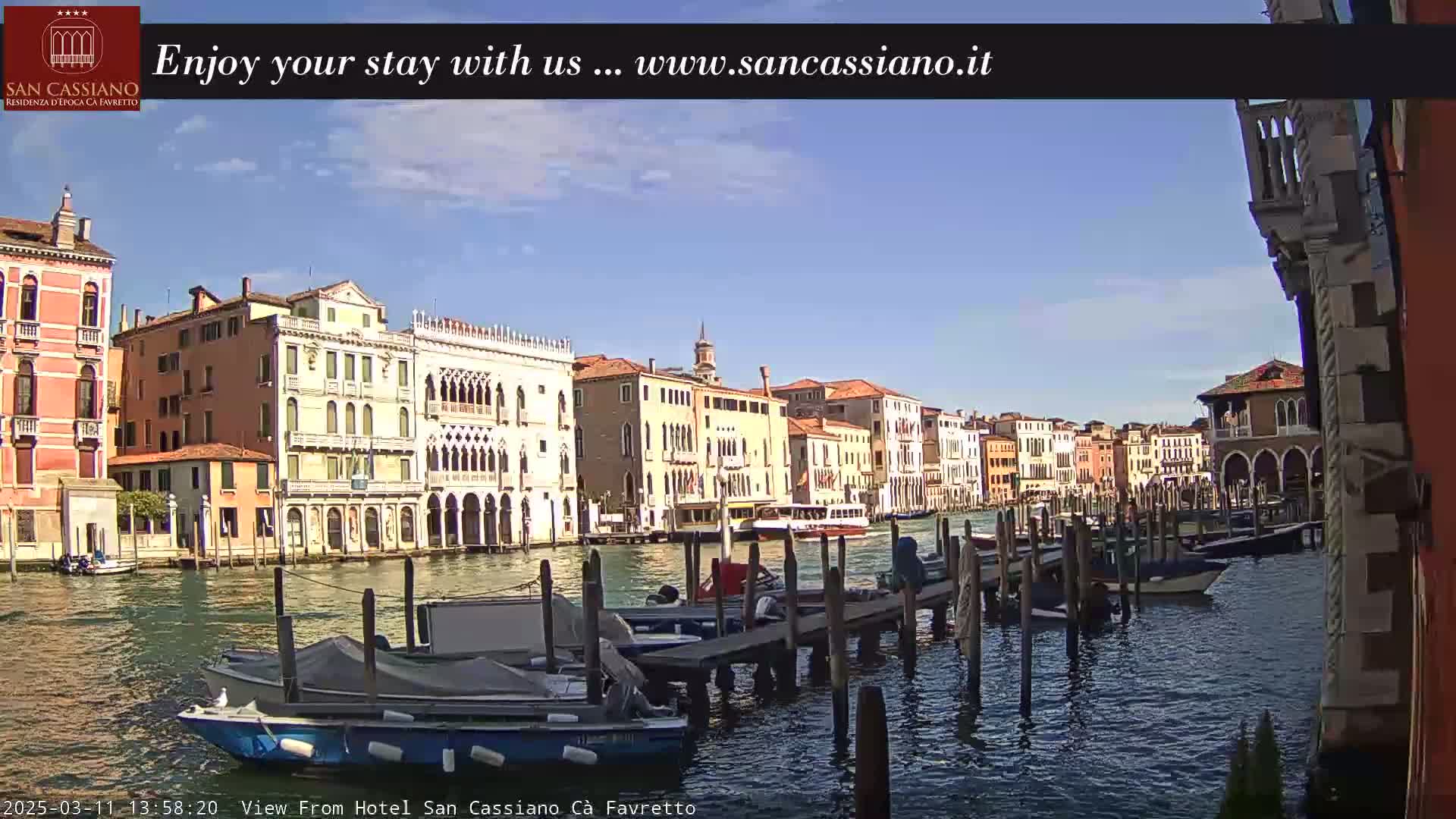 A sunny day in Venice shows a canal lined with colorful buildings, boats moored at a dock, and a larger vessel in the distance.