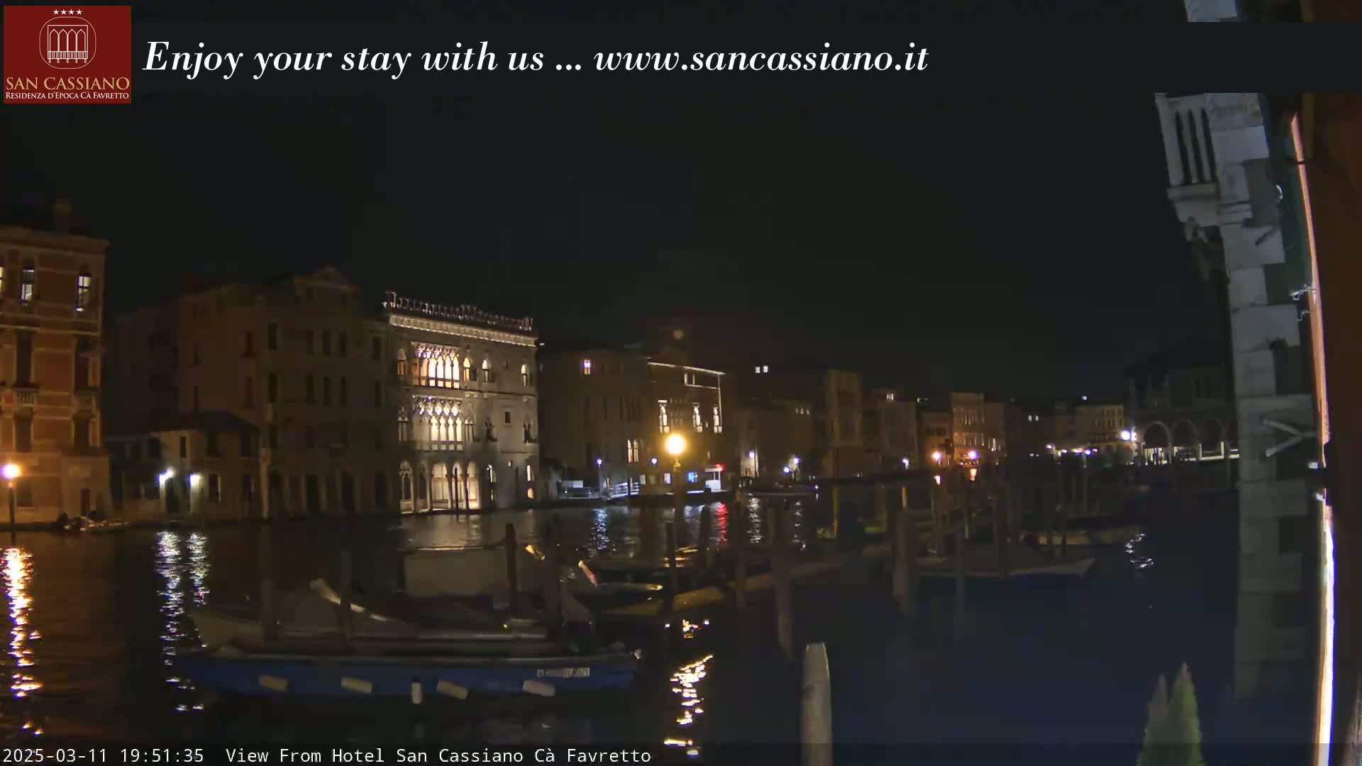 A nighttime view of a canal in Venice, Italy, showing illuminated buildings and several boats moored along the waterfront.