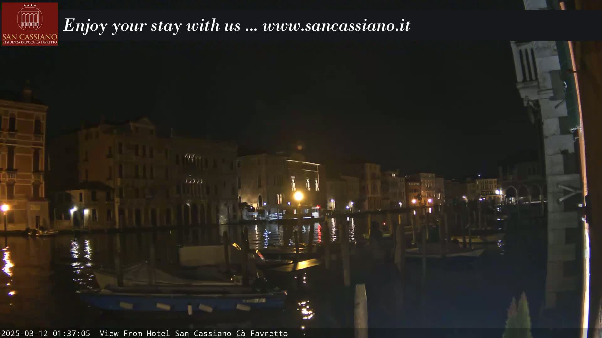 A nighttime view of a canal in Venice, Italy, shows several buildings illuminated along the waterway, with boats moored along the edge.