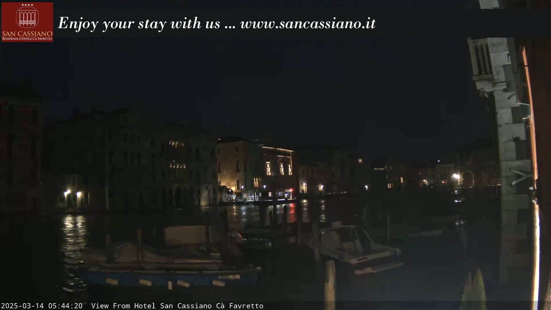 A nighttime view of a canal in Venice, with buildings dimly lit along the water's edge and boats moored nearby.