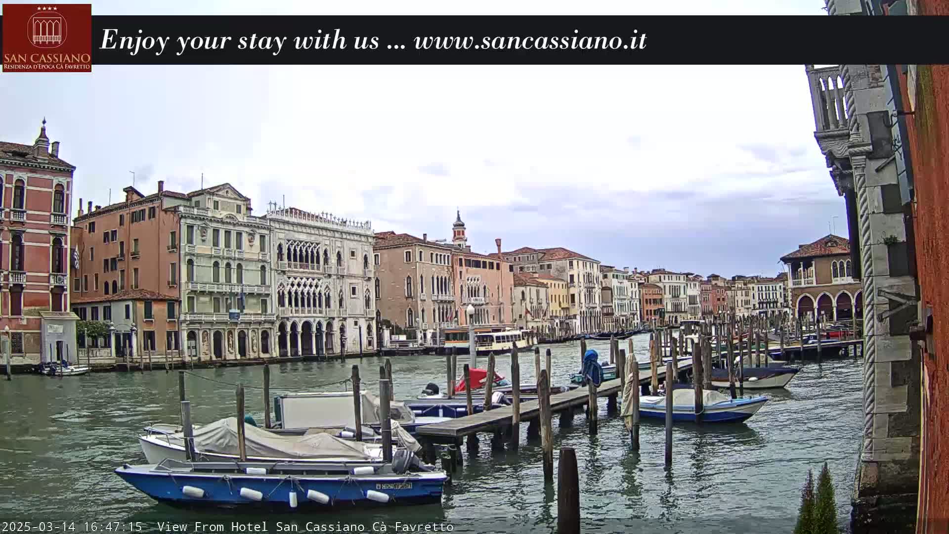 A canal in Venice, under a cloudy sky, is lined with historic buildings and numerous boats moored at wooden docks.