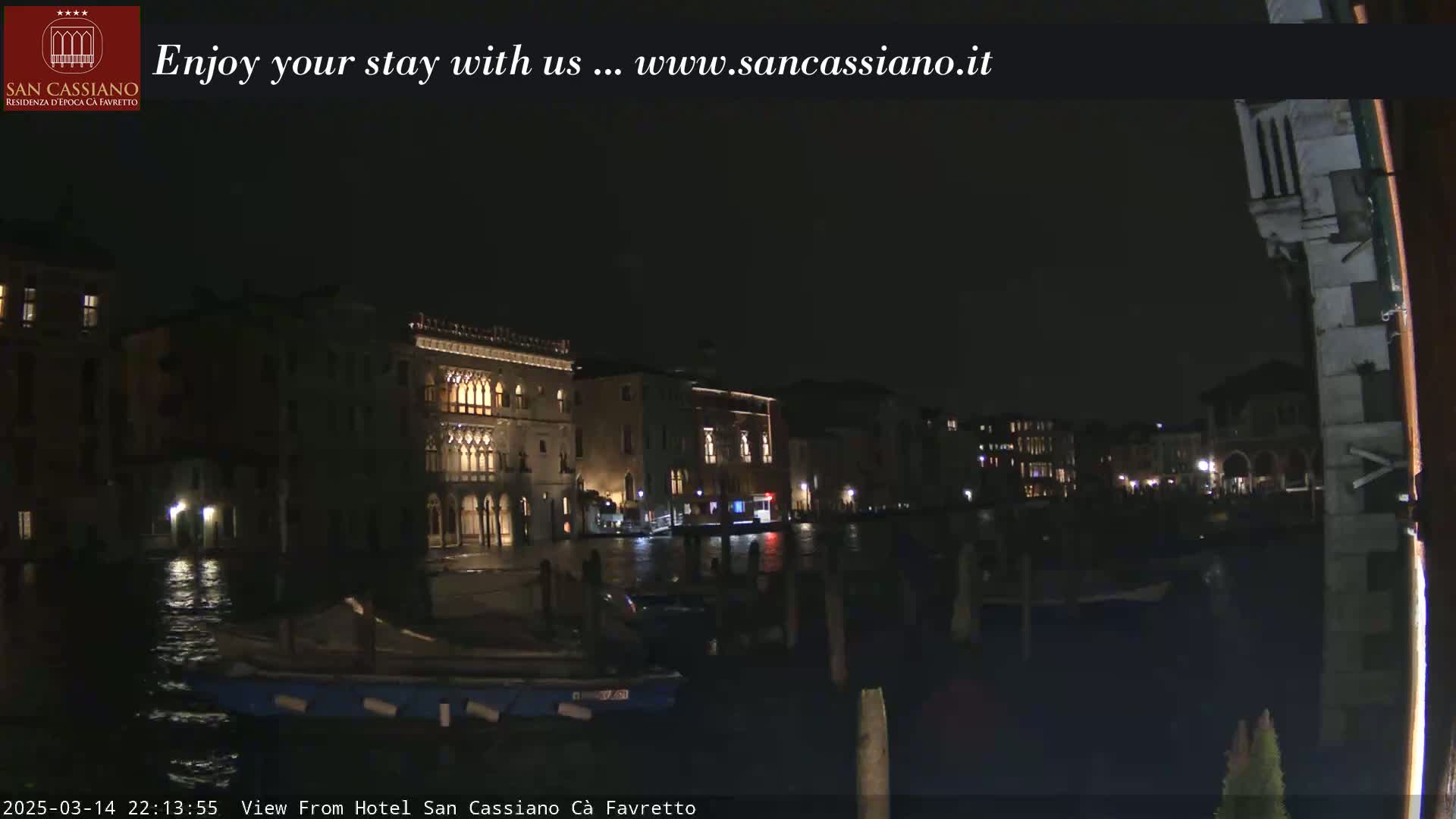A nighttime view of a canal in Venice, showing illuminated buildings along the waterway and several boats moored along the shore.