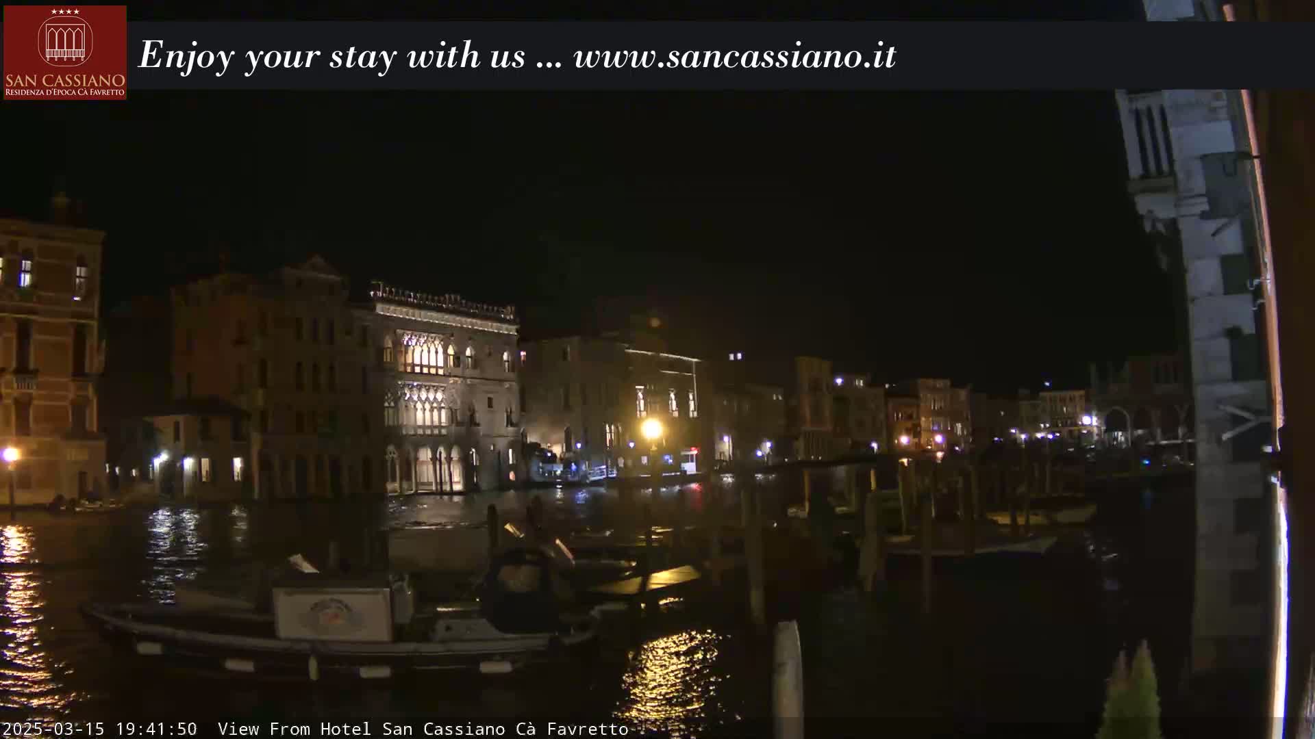 A nighttime view of a canal in Venice, Italy, shows several buildings illuminated along the waterway with boats moored along the edge.
