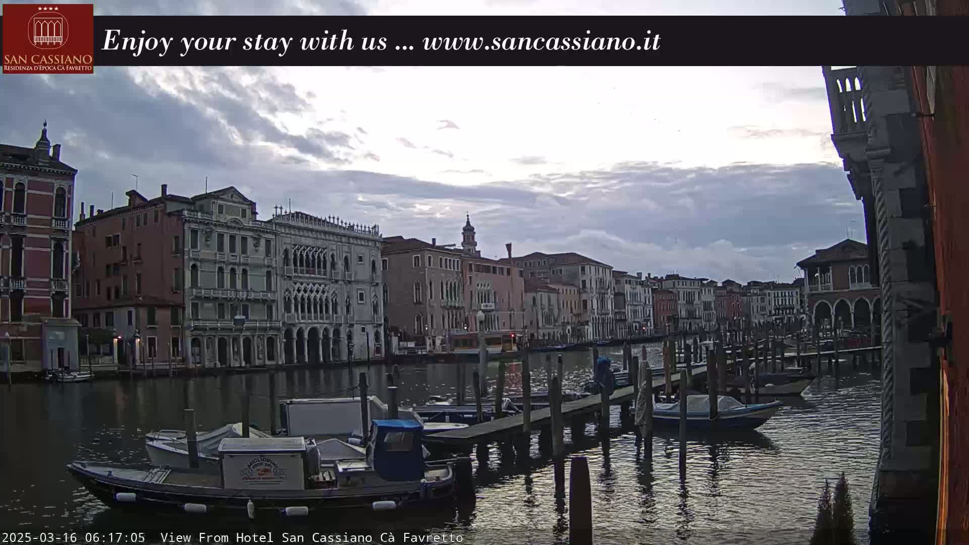 A canal in Venice, Italy, with several boats moored at a dock, under a cloudy sky.