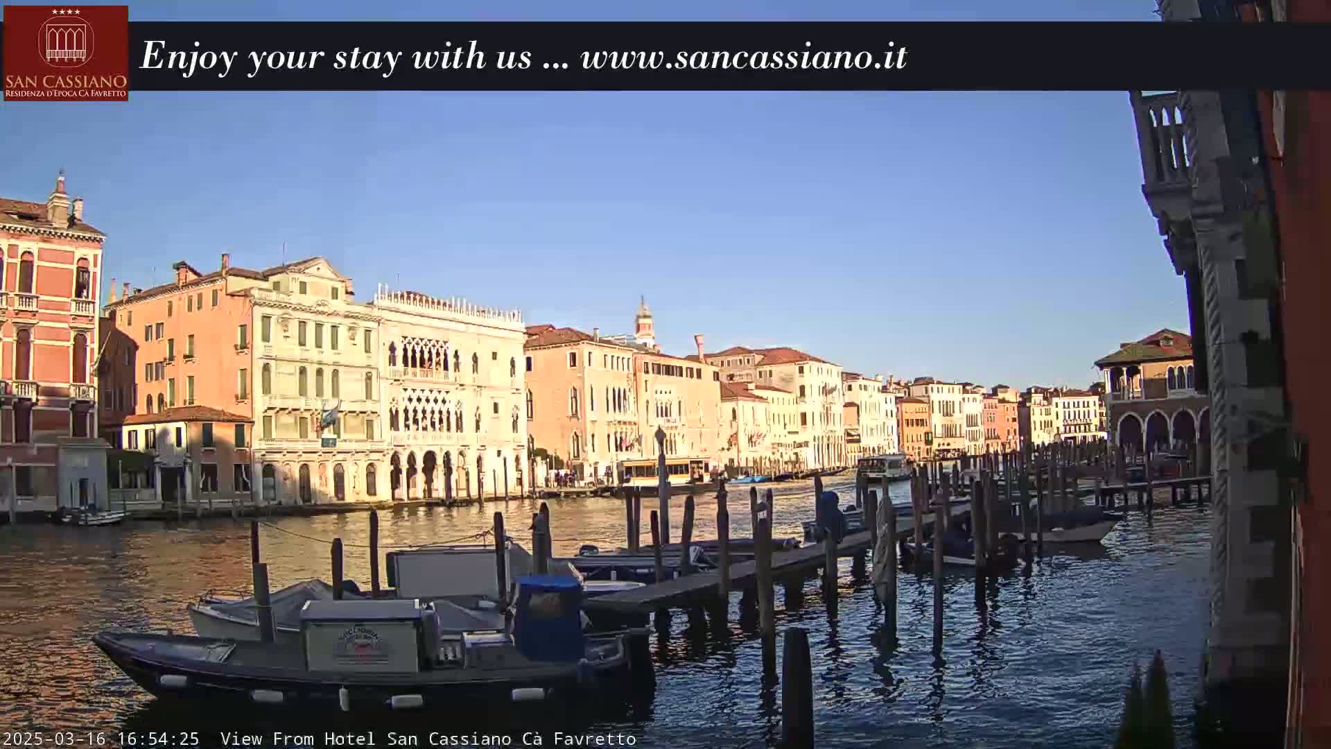 A sunny day in Venice shows colorful buildings lining a canal, with boats moored at a dock.