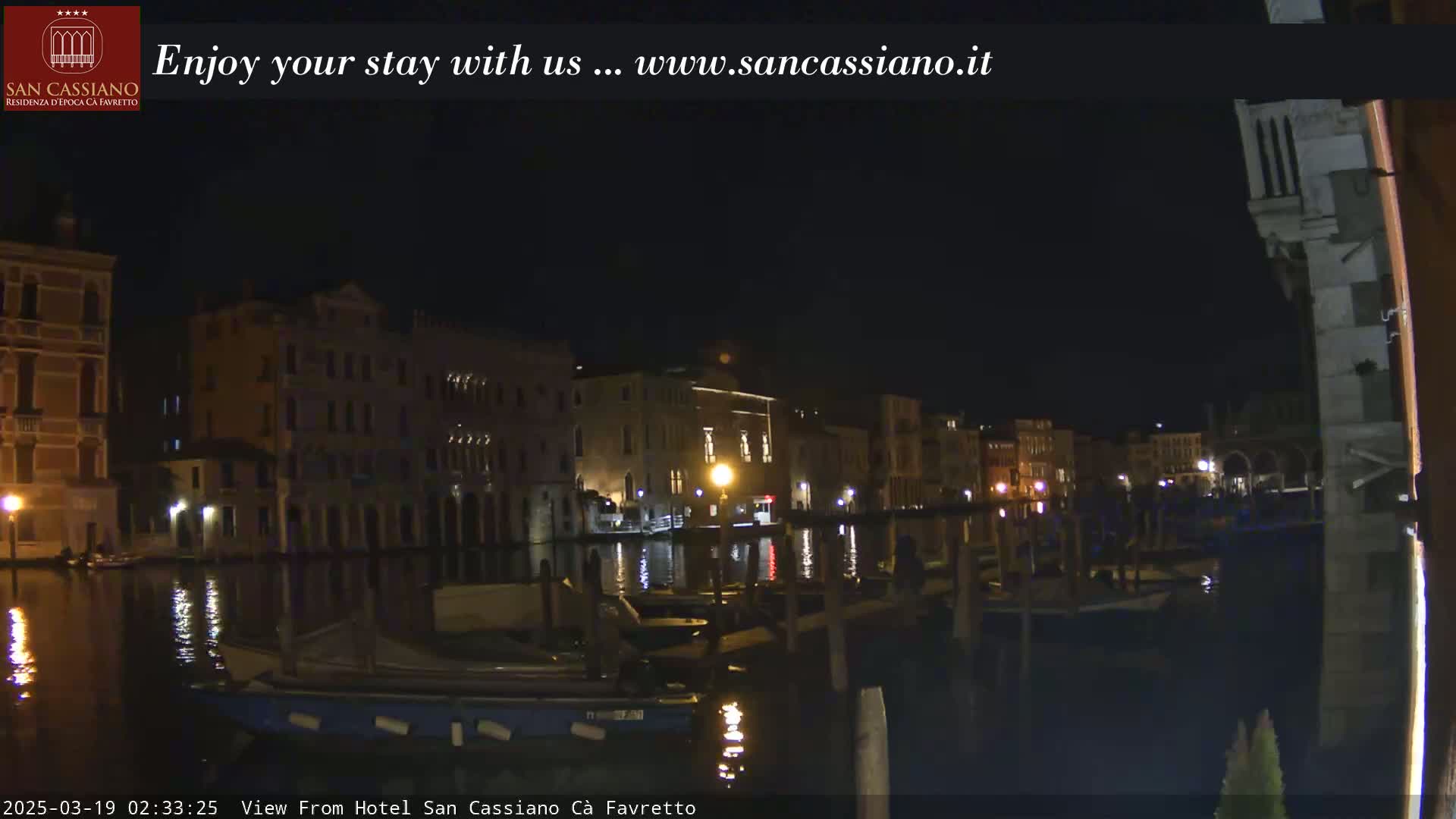 At night, several boats are moored along a canal in front of illuminated buildings.