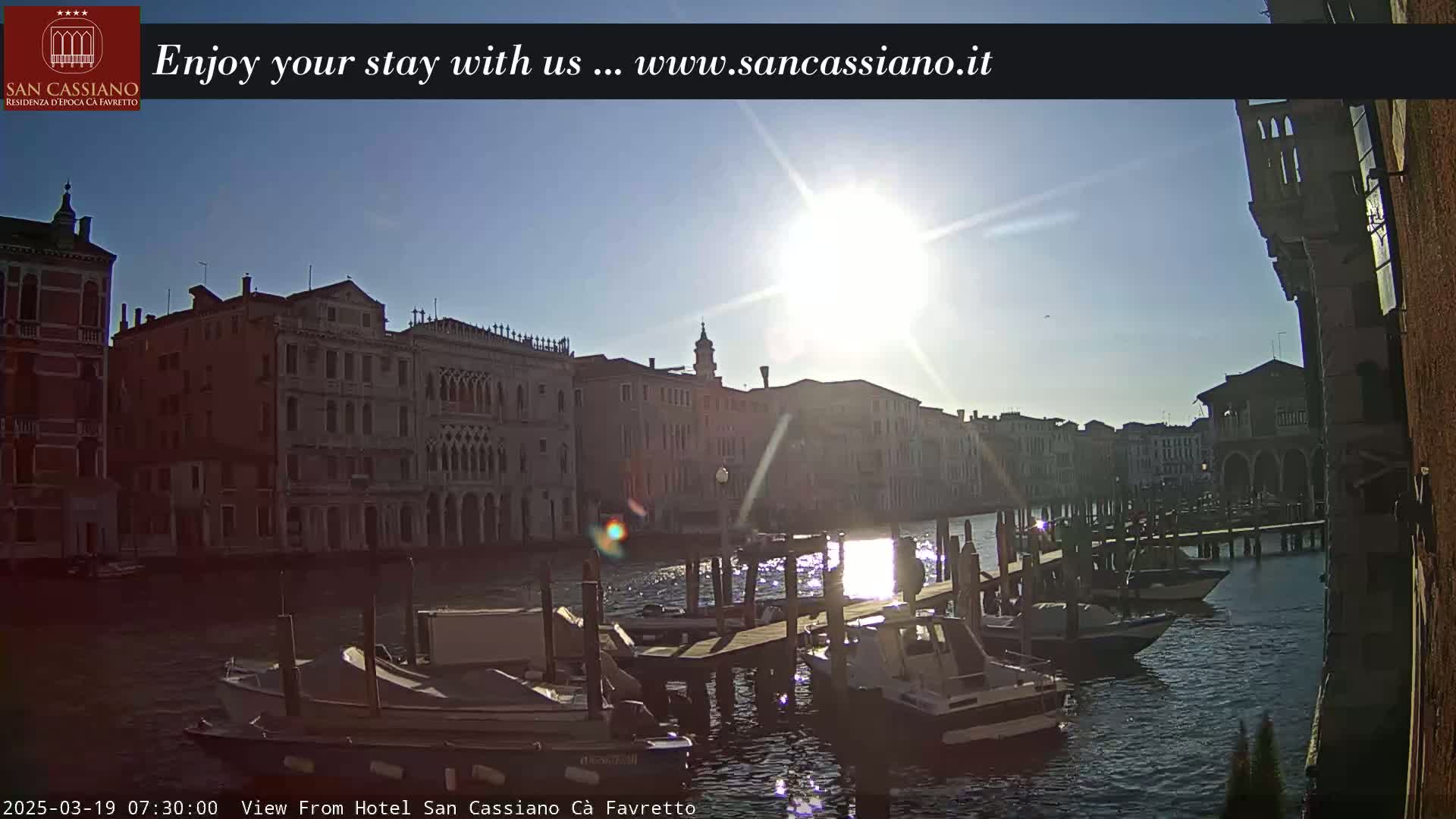 A sunny morning view of boats docked along a canal in Venice, with several buildings lining the waterway.