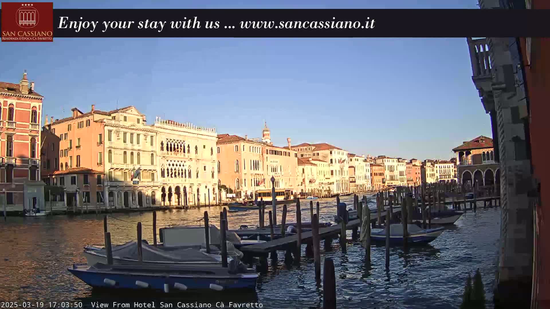 A clear, sunny day shows several canal-side buildings in Venice, Italy, with boats moored at wooden docks along a calm waterway.