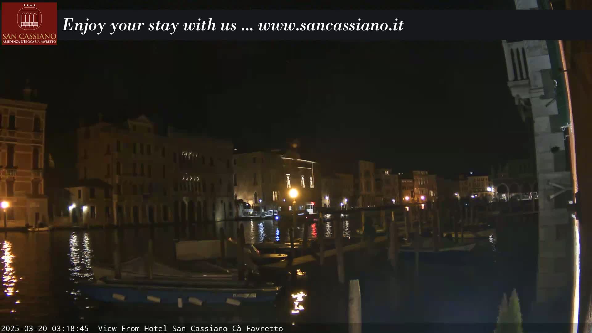A nighttime view of a canal in Venice, showing illuminated buildings along the water's edge and several boats moored at the side.