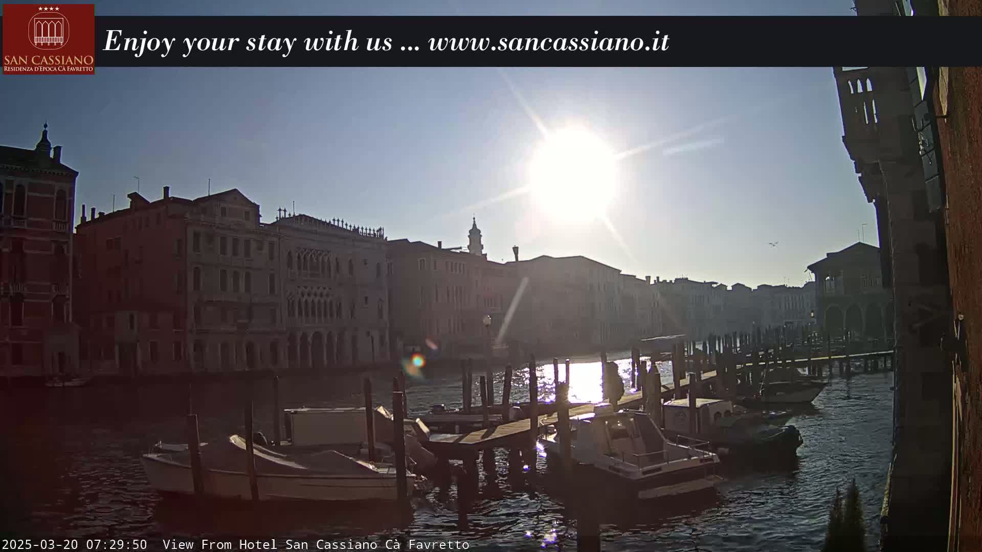 A sunny morning view of boats moored along a canal in Venice, with several buildings lining the waterway.