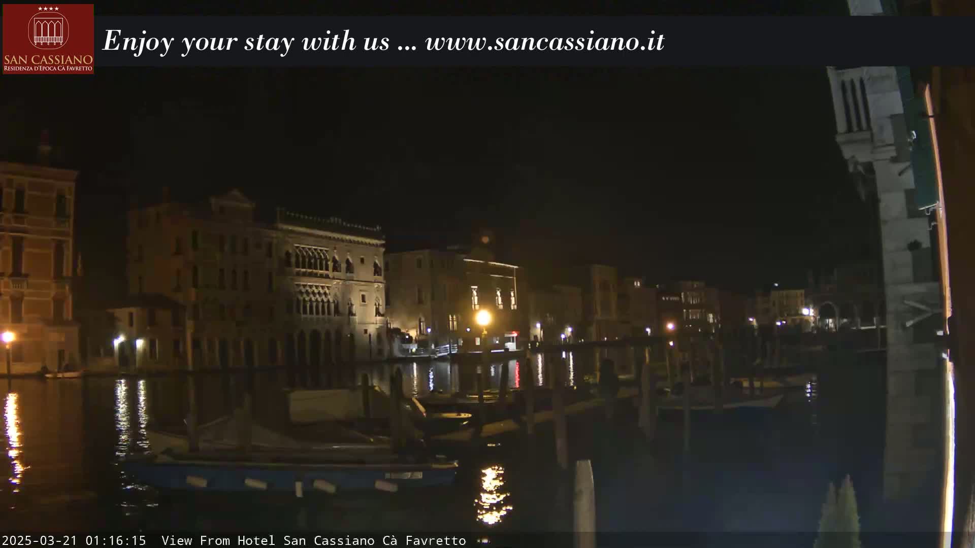 A nighttime view of a canal in Venice, showing several buildings illuminated along its banks with boats moored along the waterfront.
