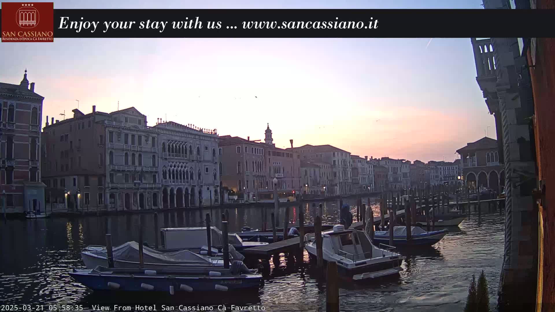 Several moored motorboats are visible on a calm canal at sunset, lined by tall, pale buildings.