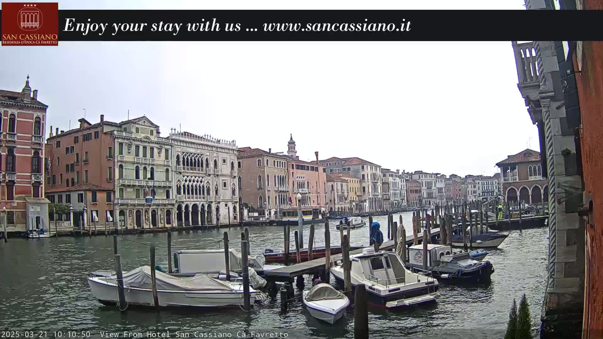 A cloudy day in Venice shows a canal lined with historic buildings and numerous boats moored at docks.
