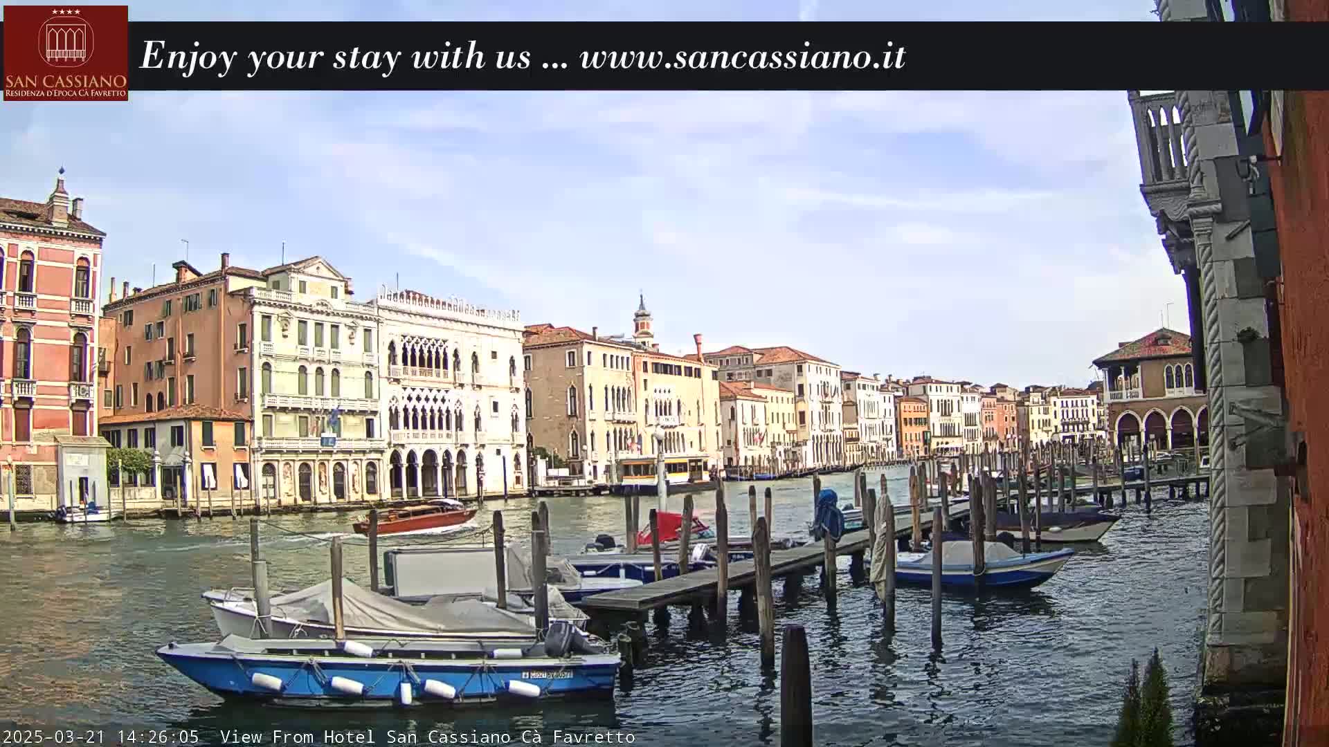 A canal in Venice, Italy, with several boats moored at a dock, under a partly sunny sky.
