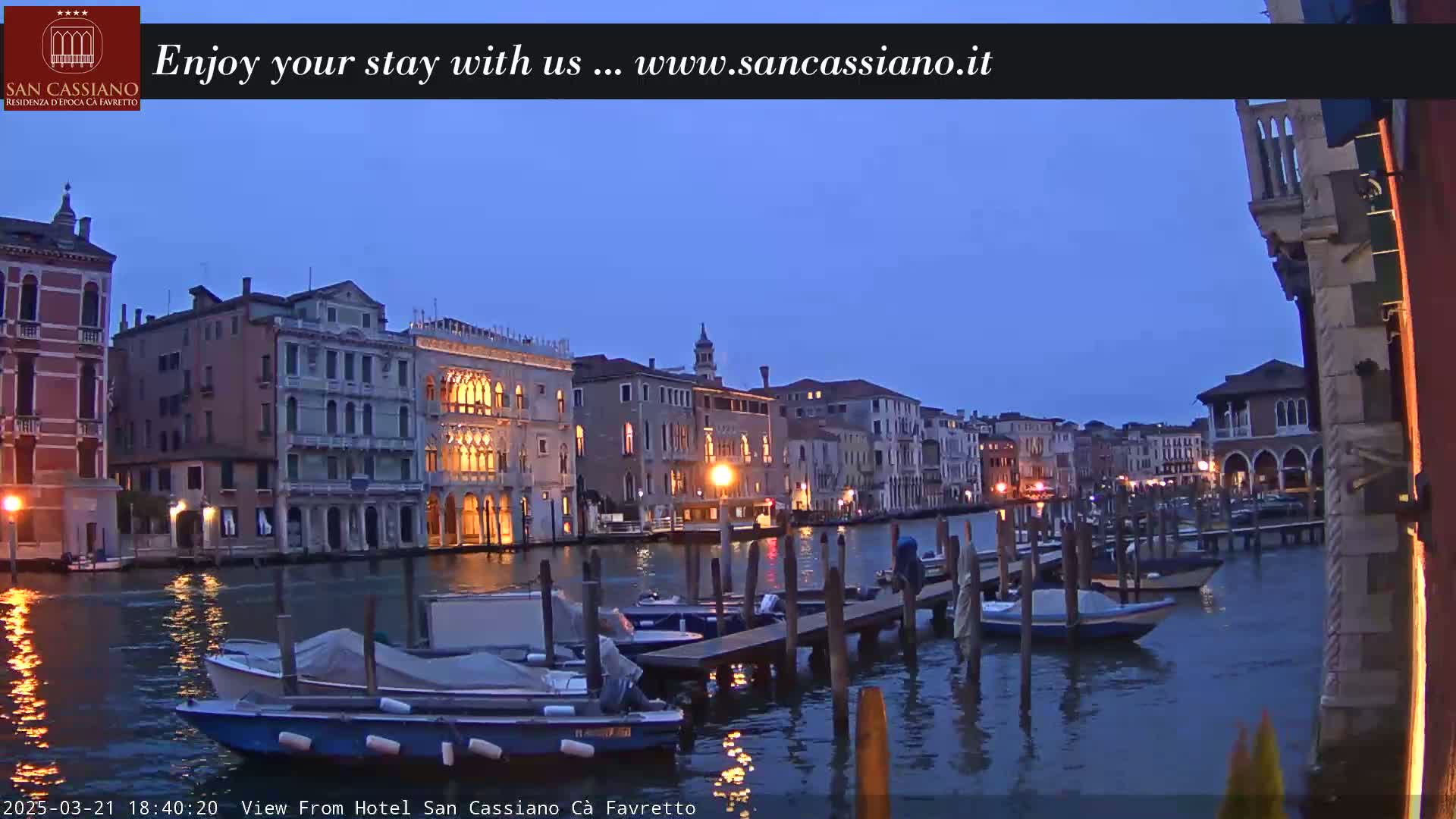 A twilight view of a Venetian canal, lined with illuminated buildings and moored boats.