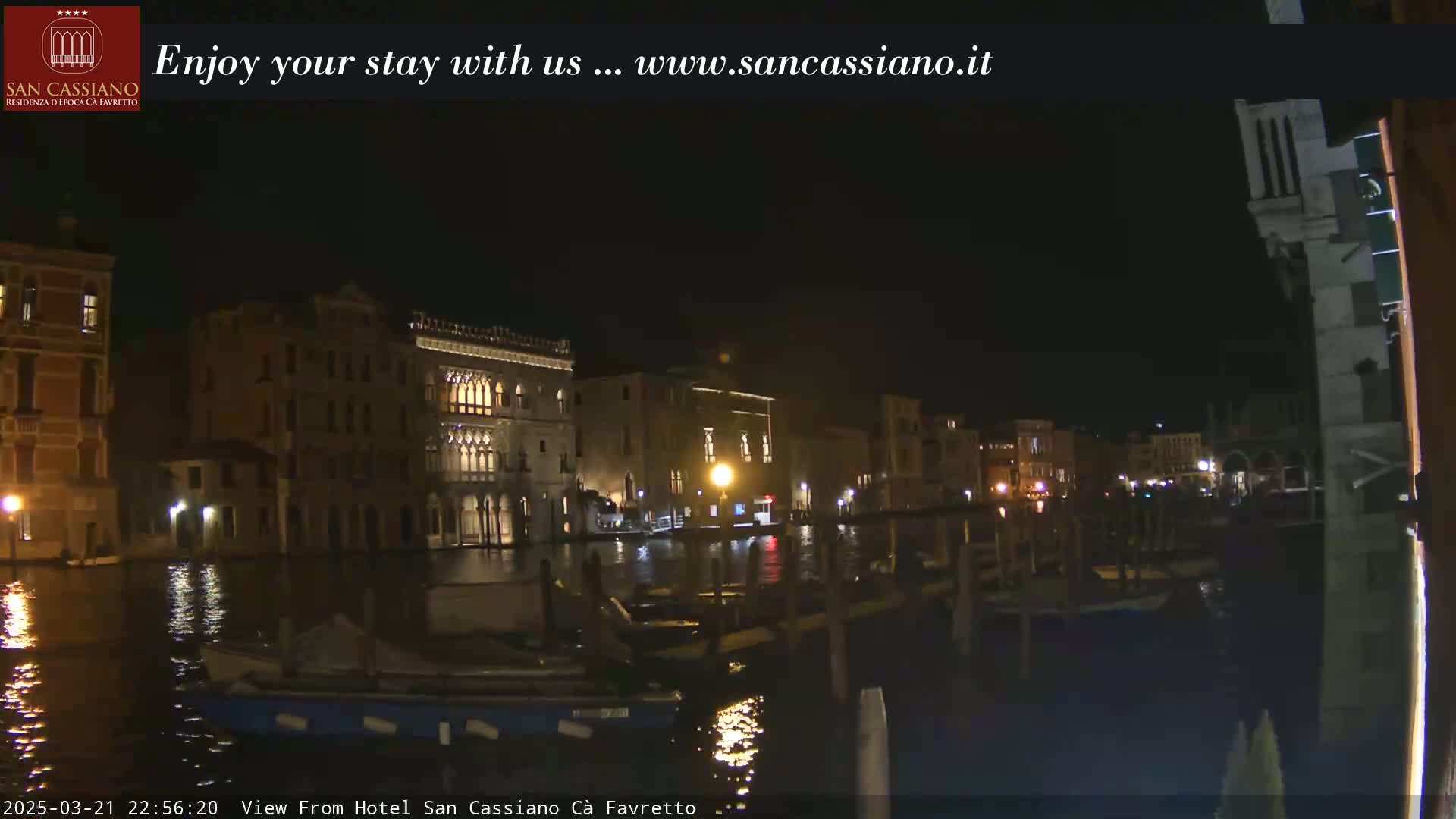 At night, illuminated buildings line a dark canal, with several boats moored along the waterfront.