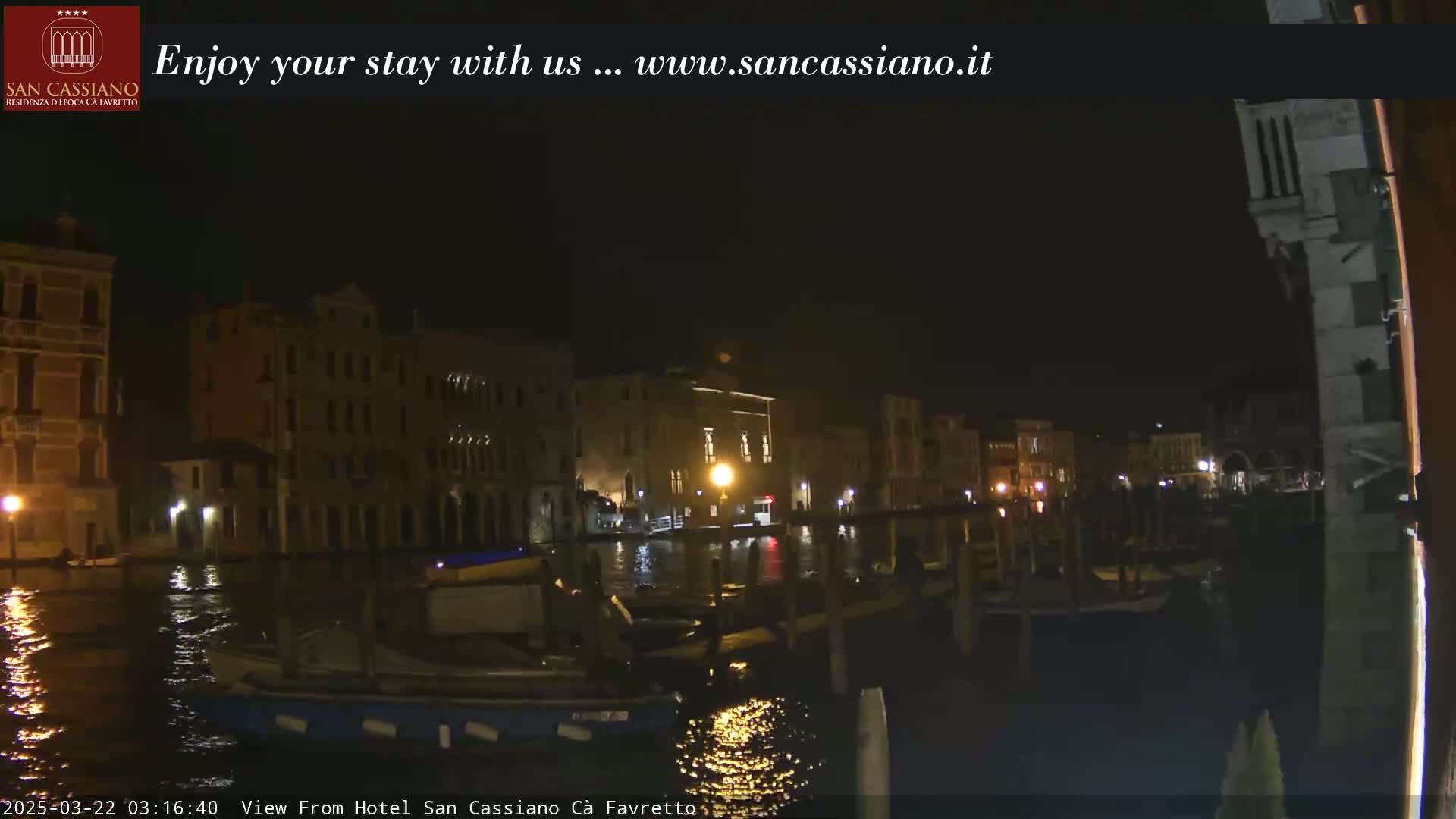 A nighttime view of a canal in Venice, with buildings lit along the waterway, and several boats moored at a dock.