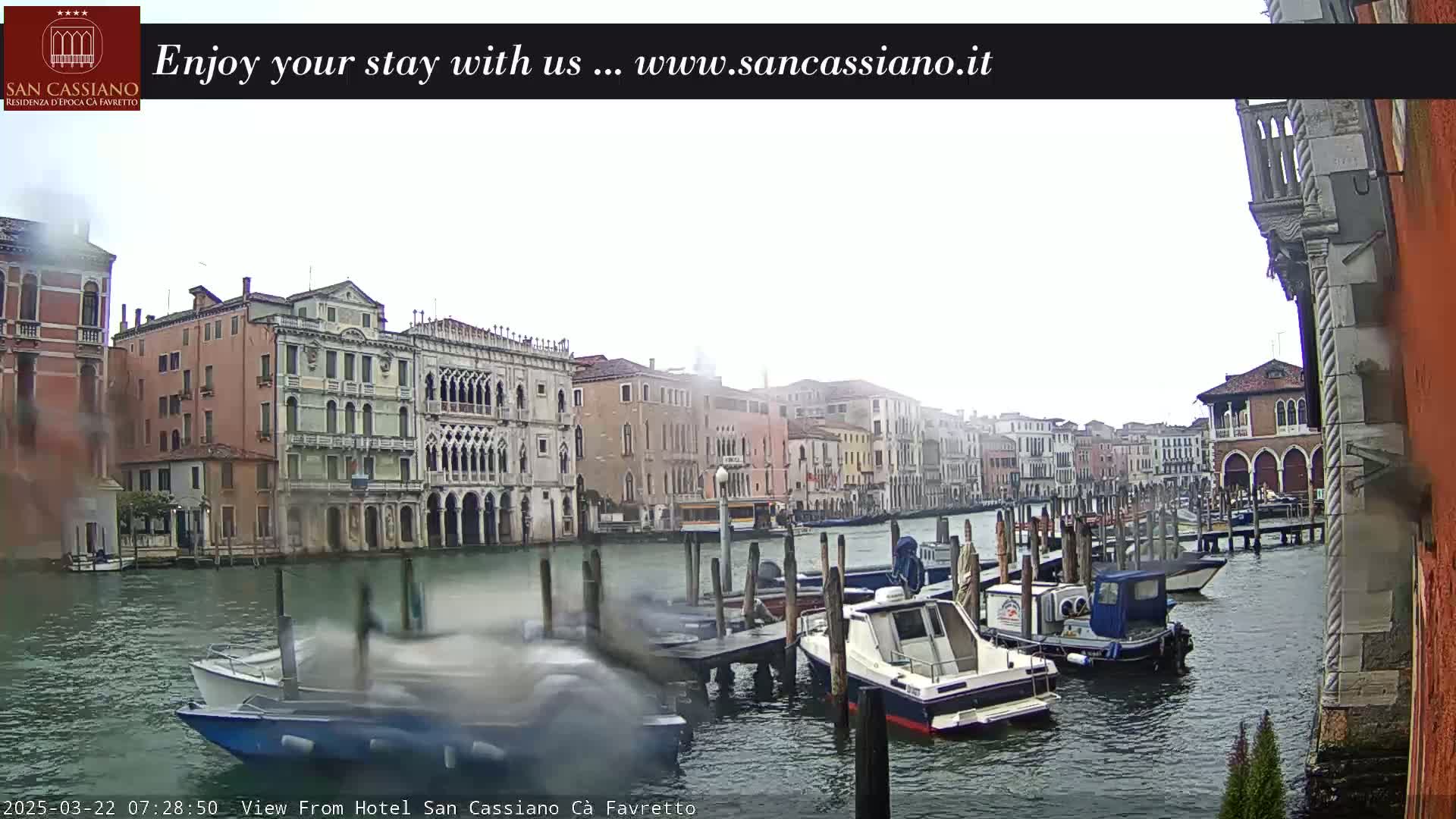 A canal in Venice, Italy, is shown on an overcast day with several boats moored at the side, and buildings lining the waterway.
