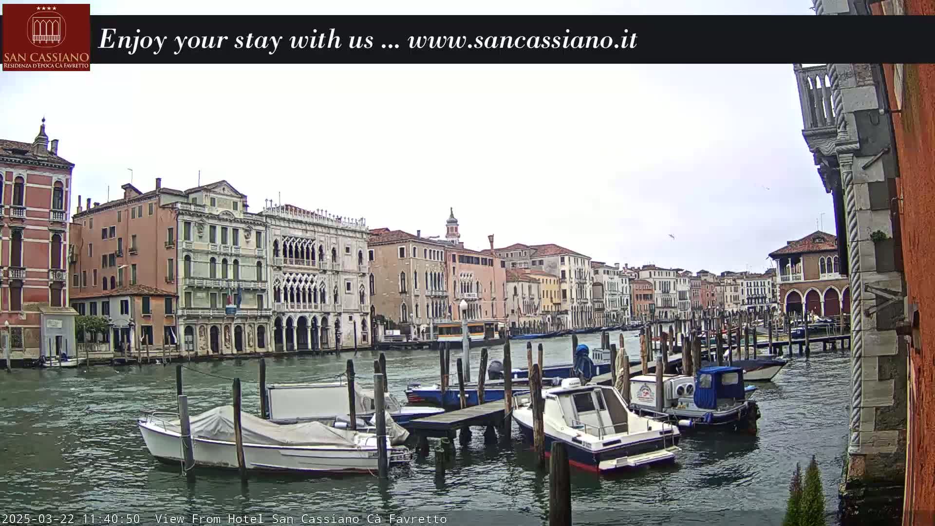Several boats are moored along a canal lined with tall, historic buildings under an overcast sky.