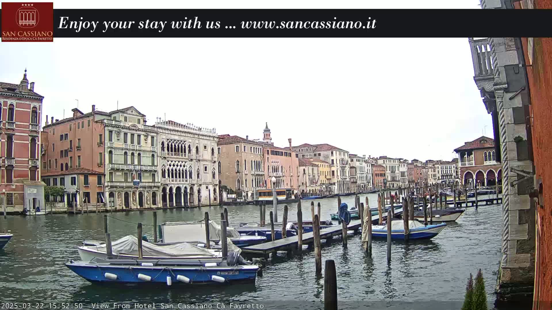 A canal in Venice, Italy, with numerous boats moored along wooden docks, is viewed from a building on an overcast day.