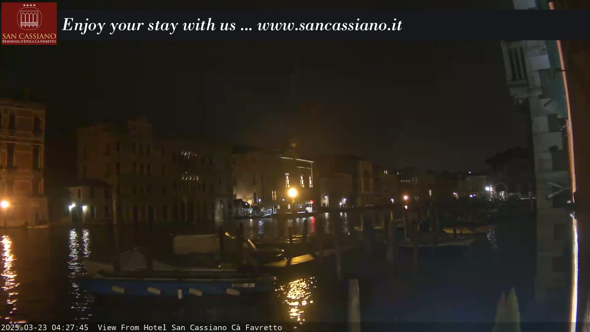 A nighttime view of a canal in Venice, Italy, showing dark buildings and boats along the water's edge under a clear, dark sky.
